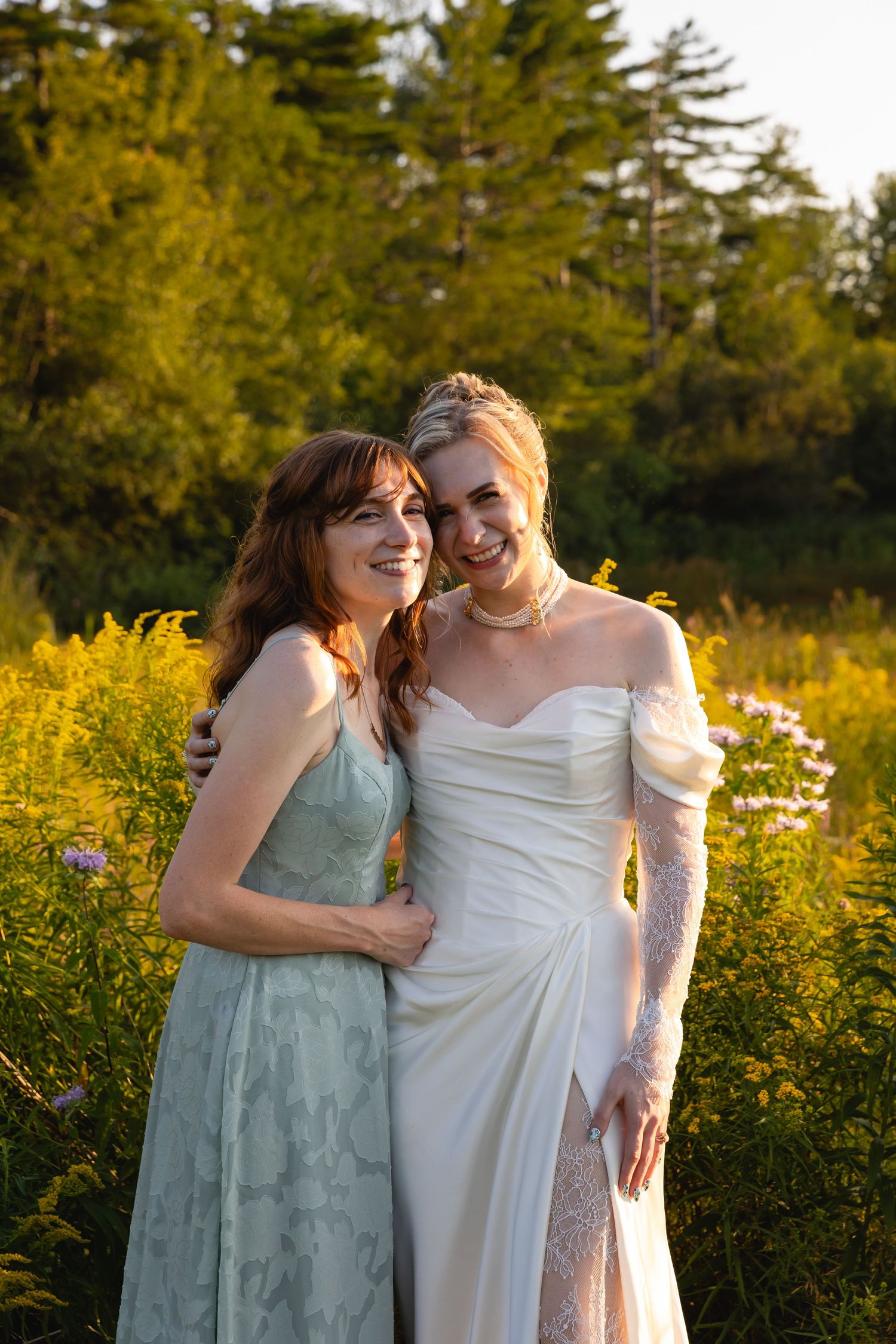 Two people in elegant dresses share a joyful moment together in a sunlit meadow with wildflowers.