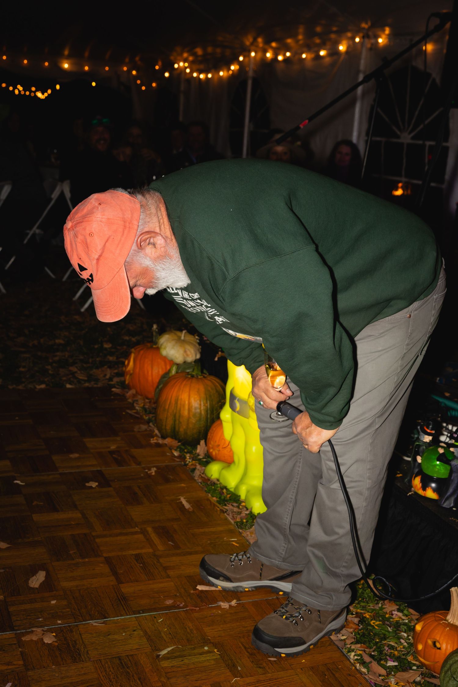 A person in a green jacket and orange hat bends down to inspect Halloween pumpkins under string lights at night.