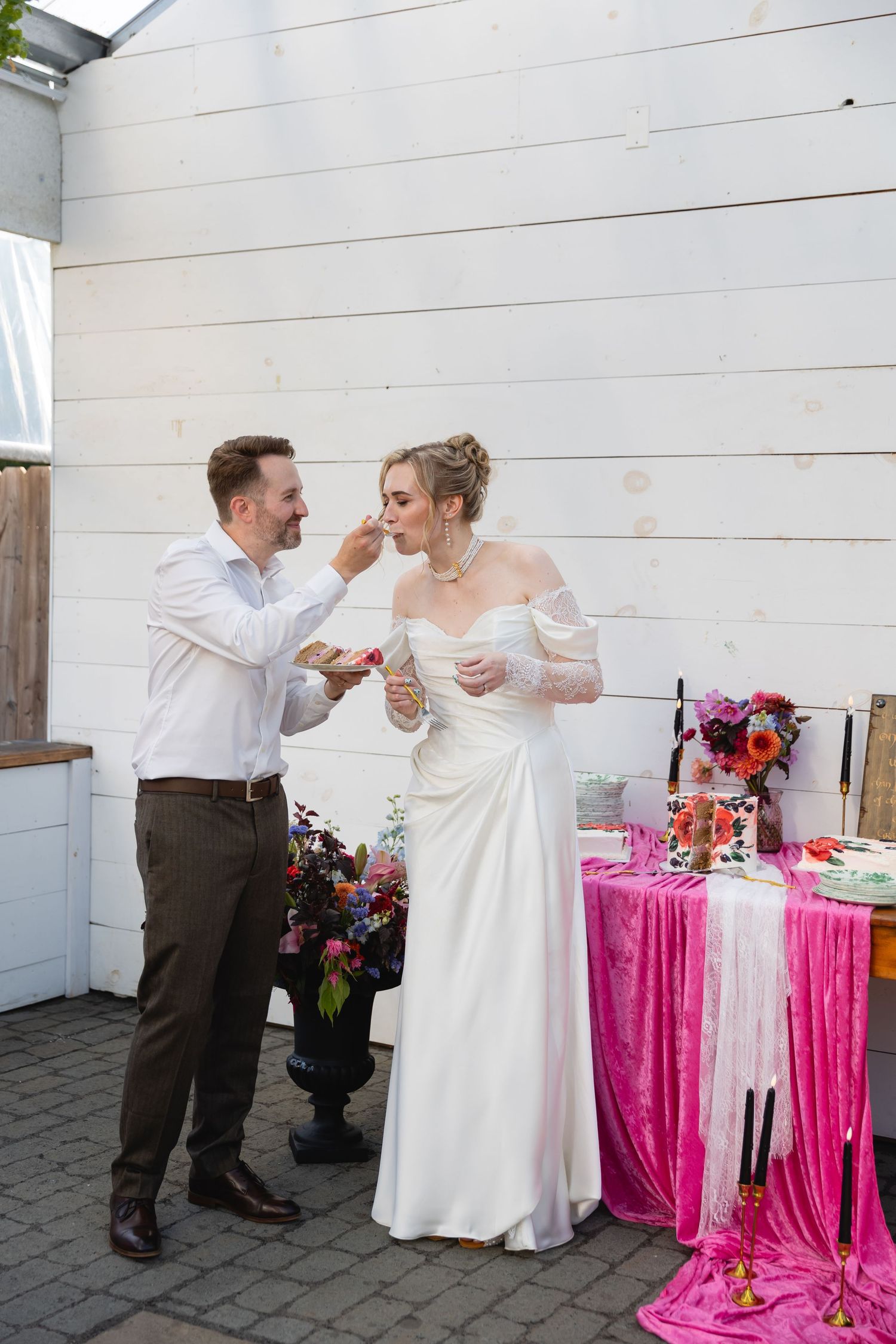 Wedding couple shares playful cake-feeding moment at reception with bright pink tablecloth and floral decorations.