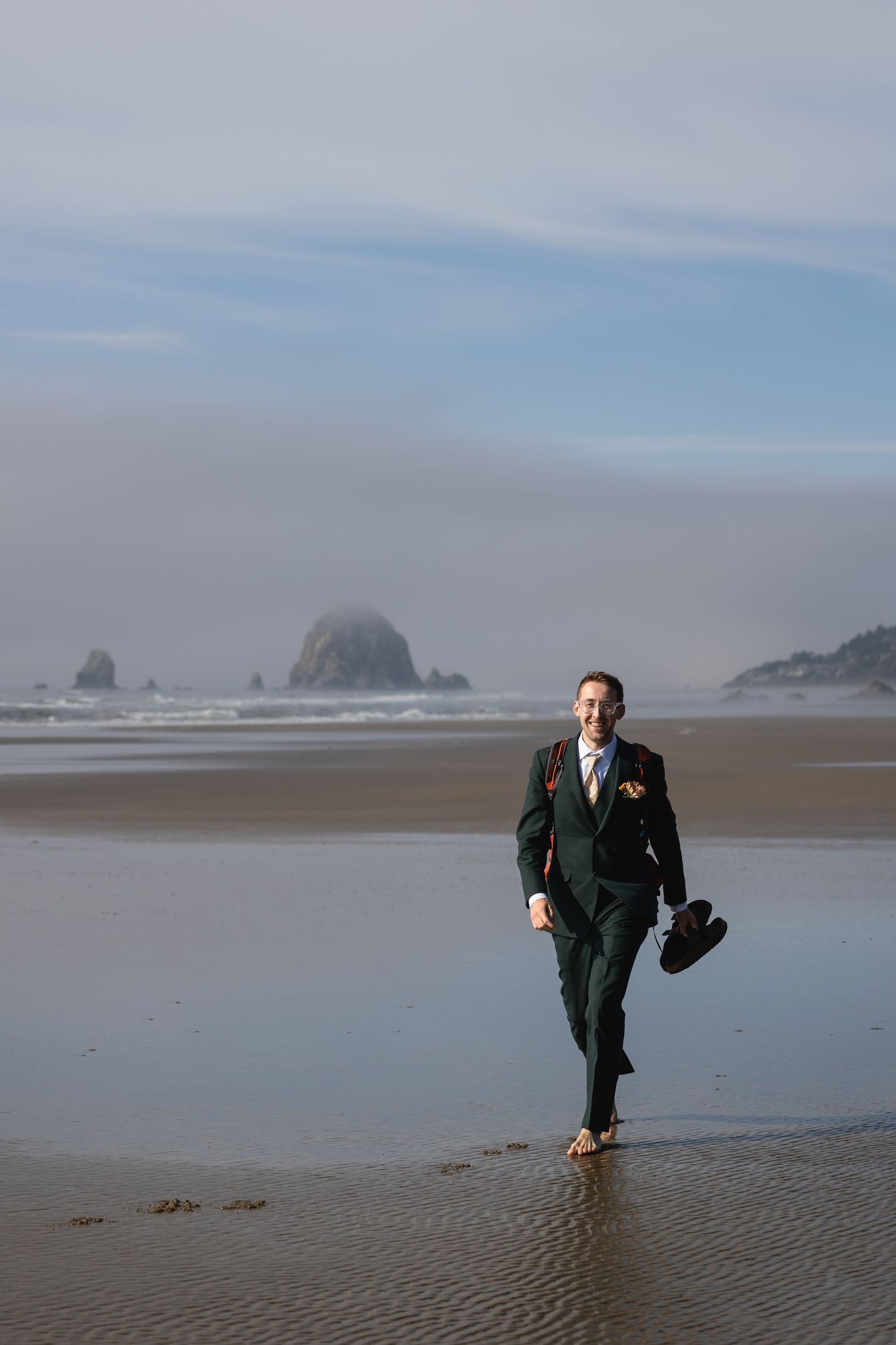 A person in a dark suit walks along a misty beach with coastal rock formations visible in the background.