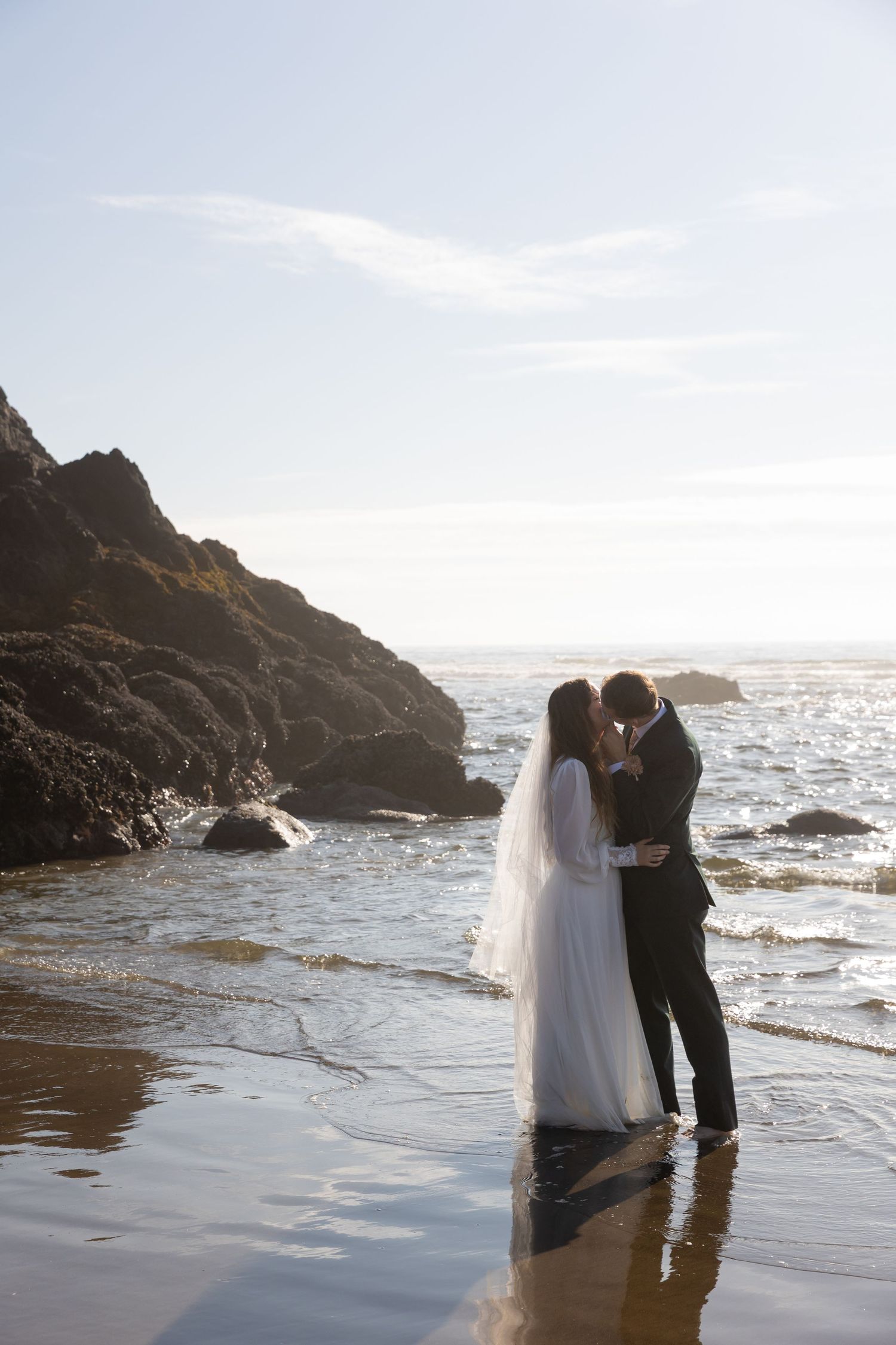 A couple shares an intimate moment on a rocky beach at sunset with waves crashing against the shore.