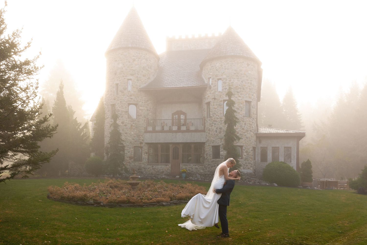 Wedding couple share a romantic moment on a green lawn before a fog-shrouded stone castle venue.