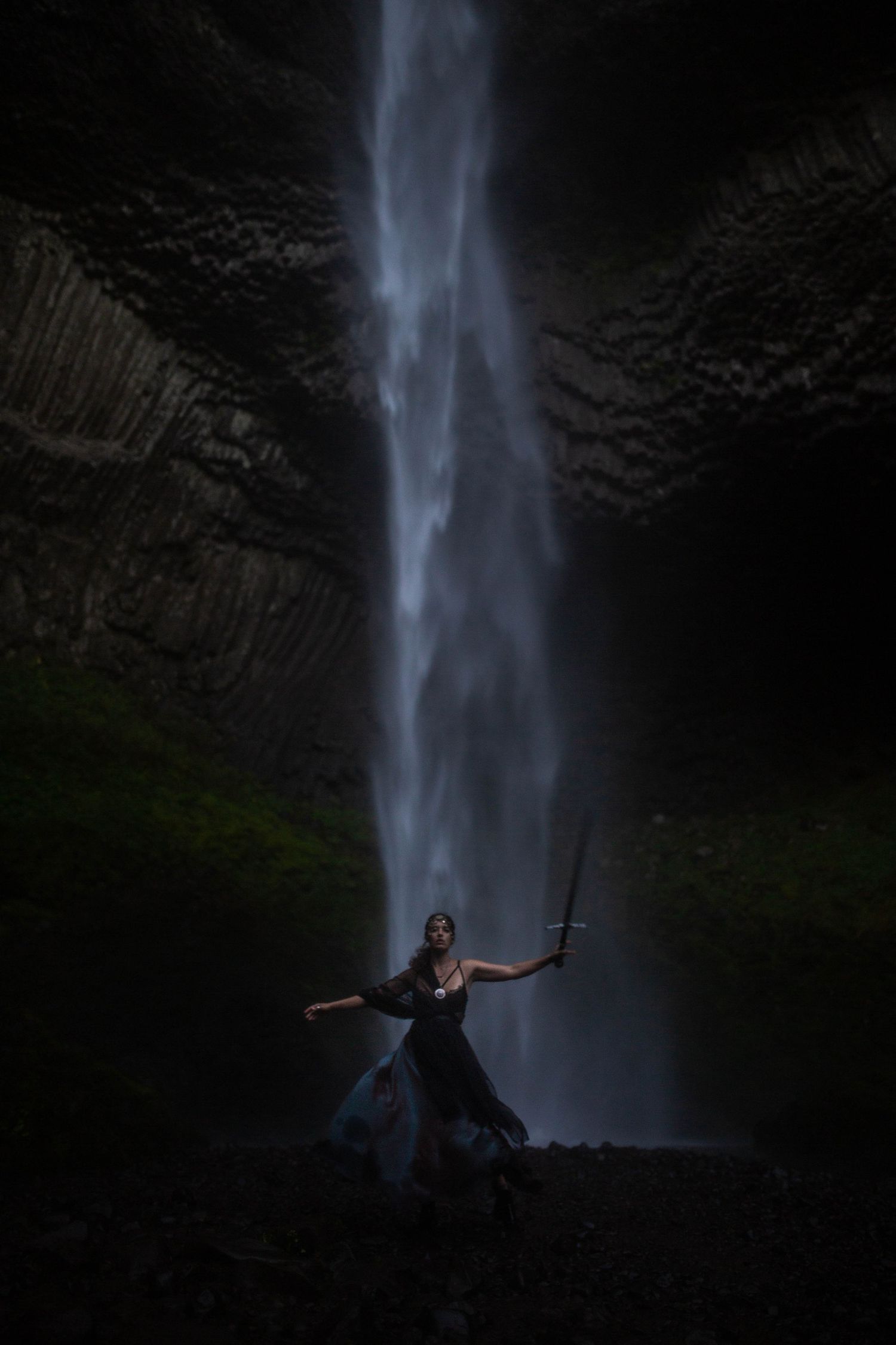 Woman in flowing dark gown wielding sword before dramatic waterfall in dark cave setting.