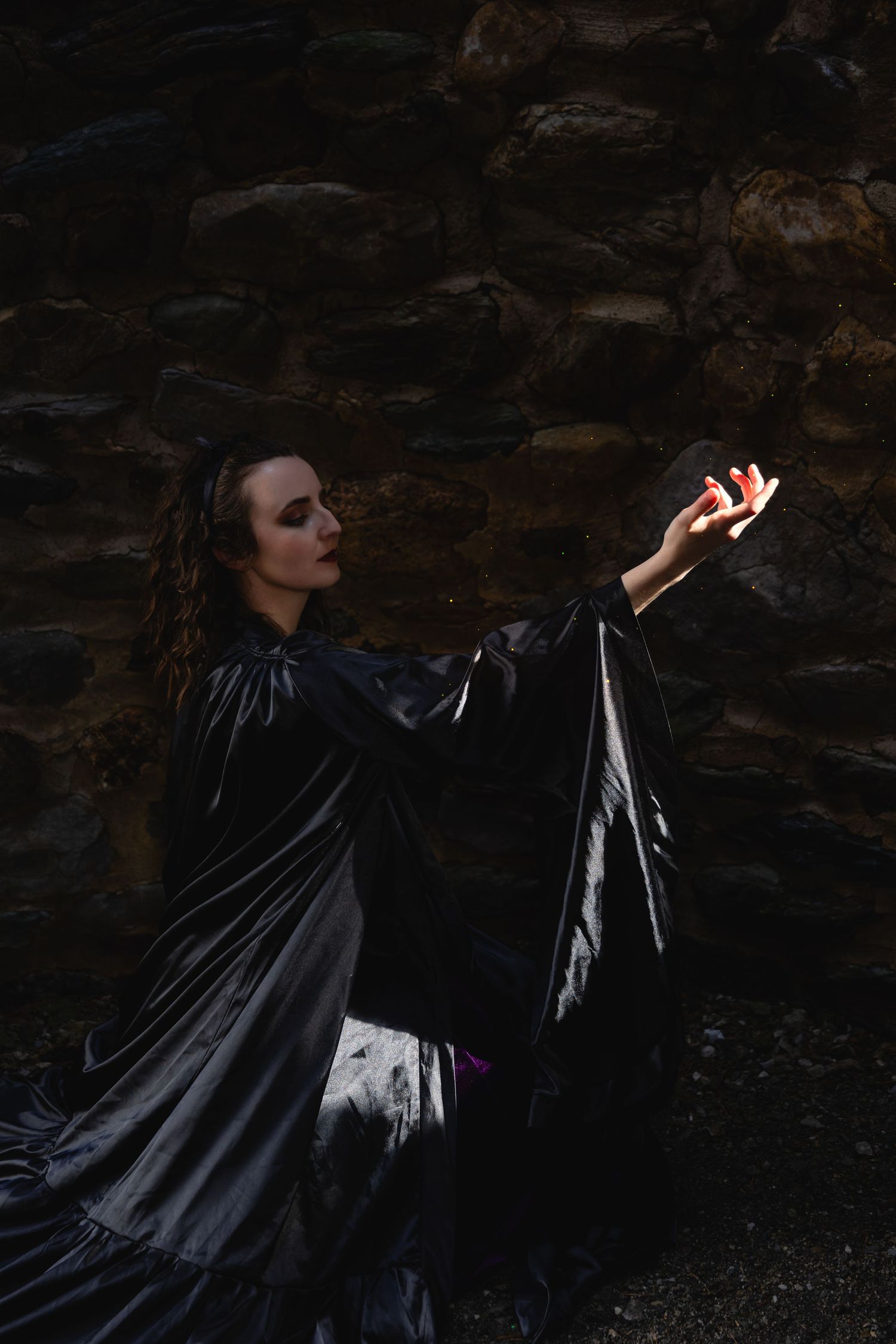 A woman in a black satin cloak reaches toward glowing light against a dark stone wall.