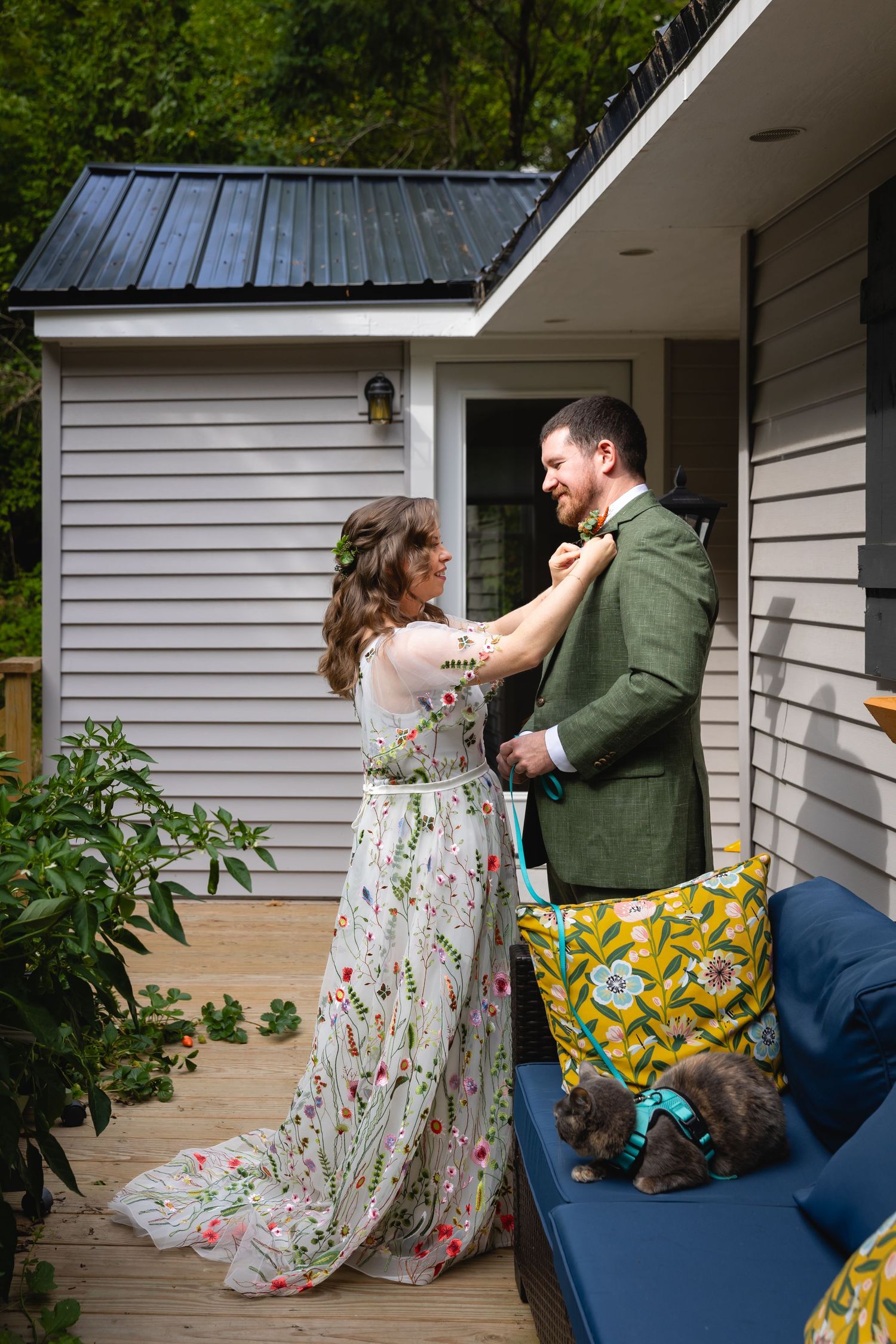 A couple in casual attire playfully interact on a porch with white siding and greenery visible in the background.