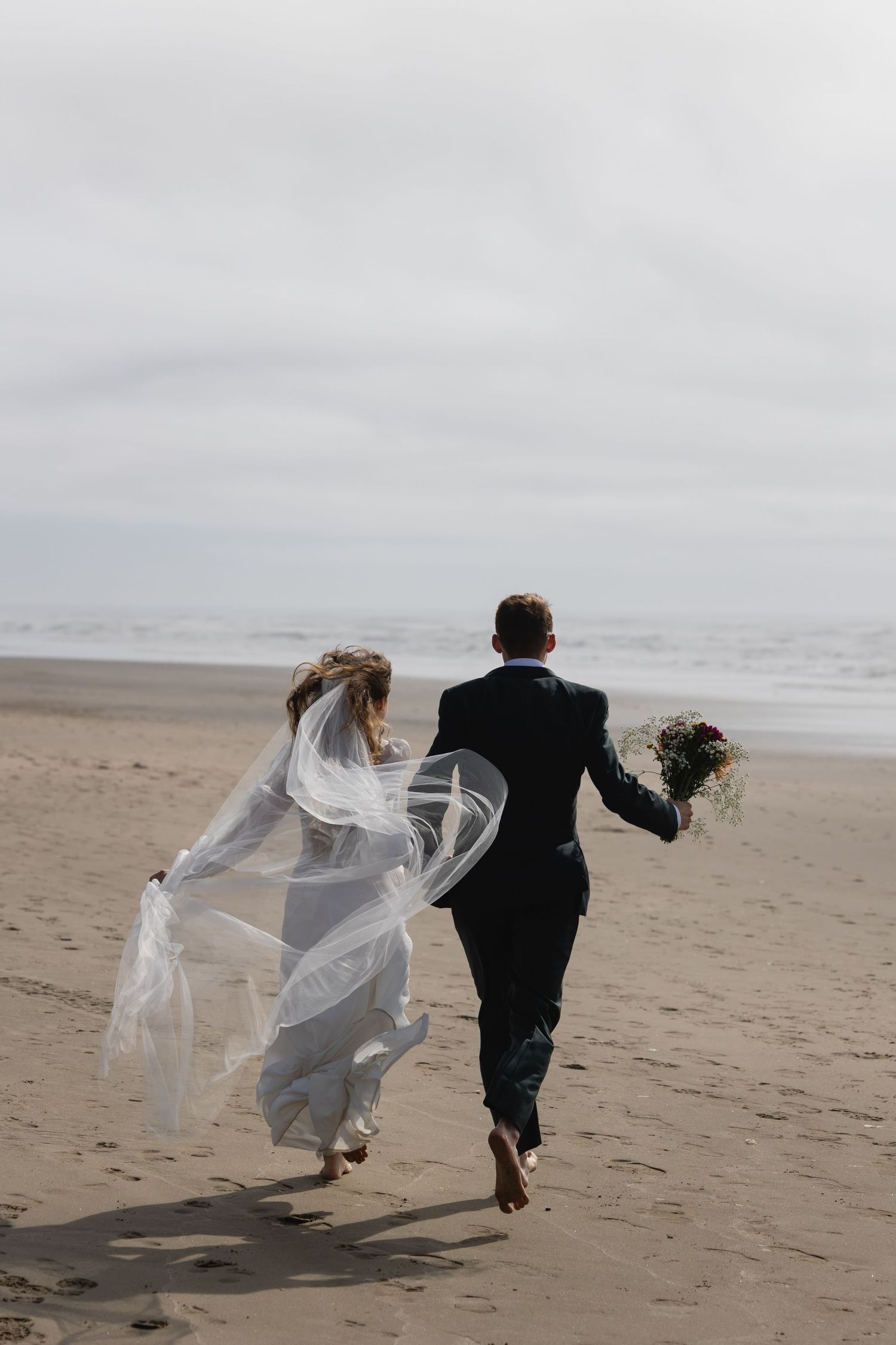 A couple in wedding attire runs playfully along a misty beach as the bride's veil flows dramatically in the wind.