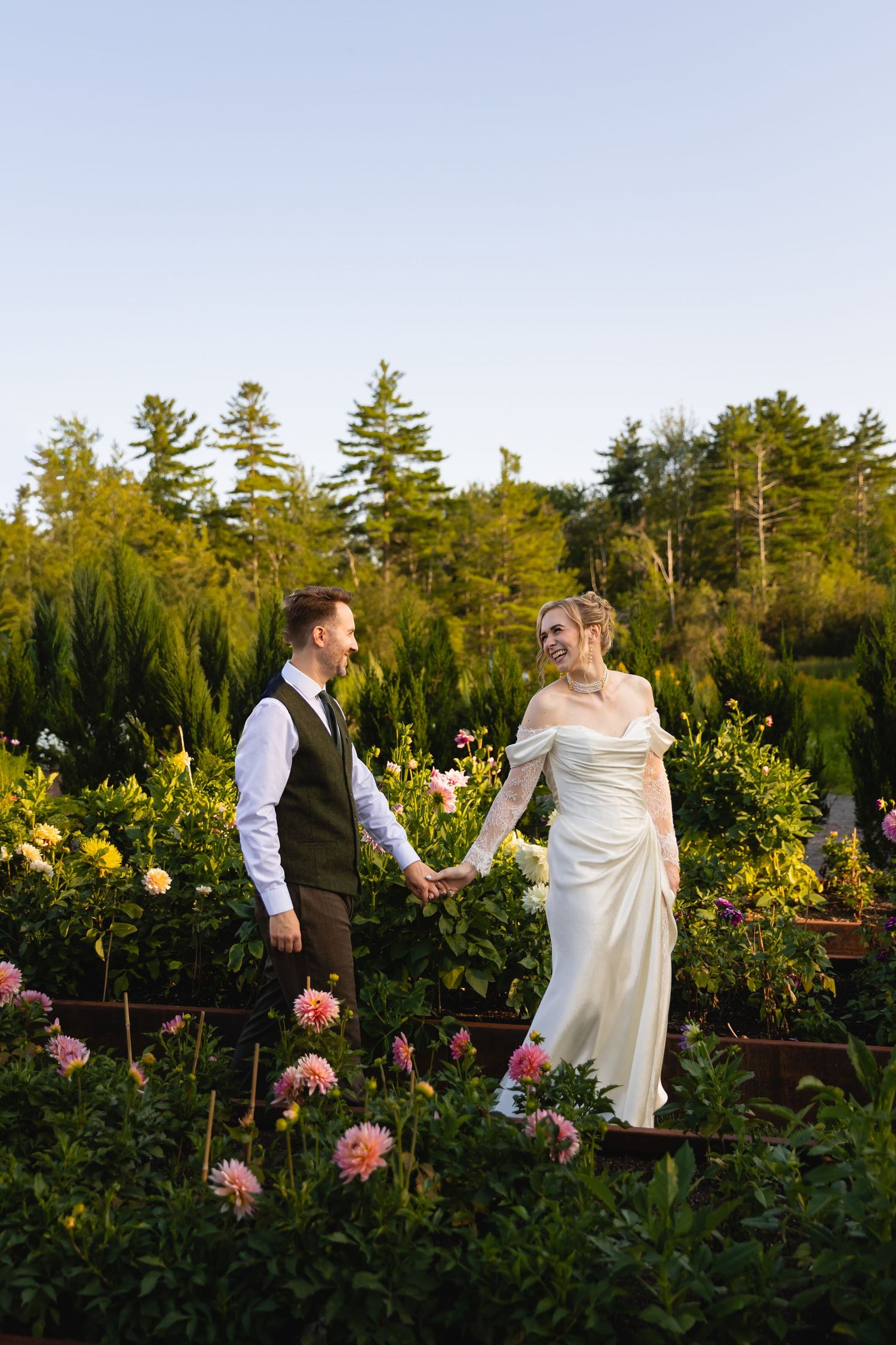 Couple in wedding attire walks together through a garden with blooming flowers at sunset among tall pine trees.