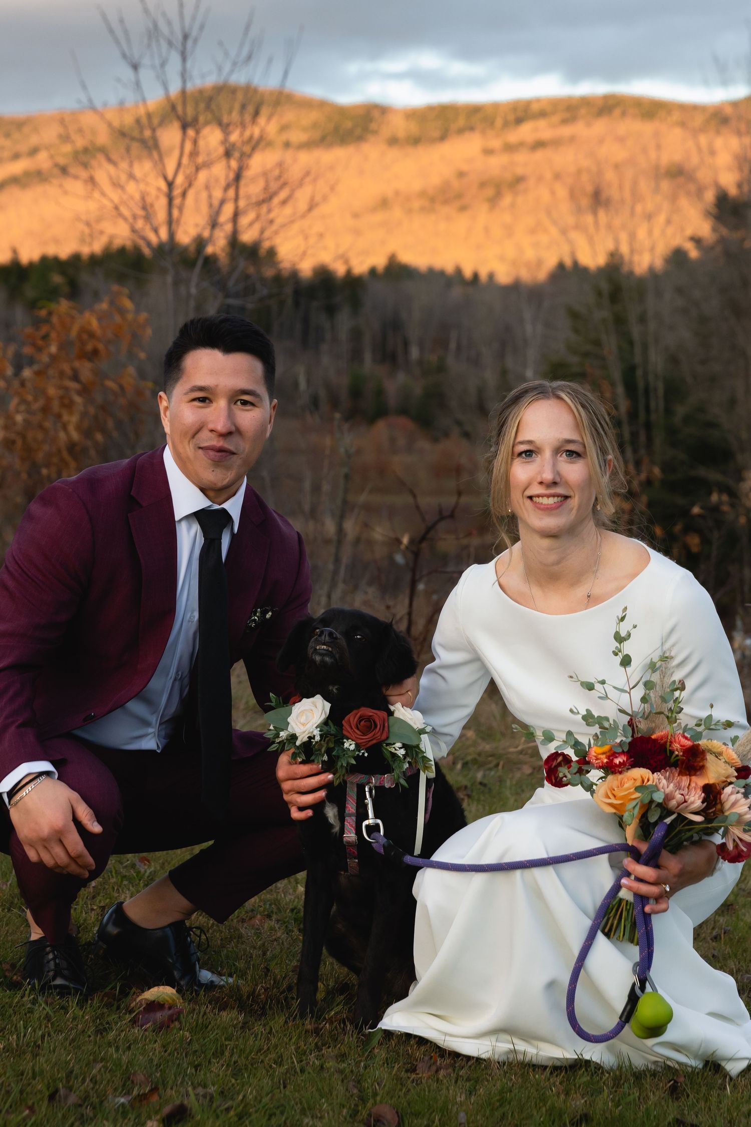 A couple in wedding attire kneels outdoors with their black dog against a backdrop of autumn mountains at sunset.