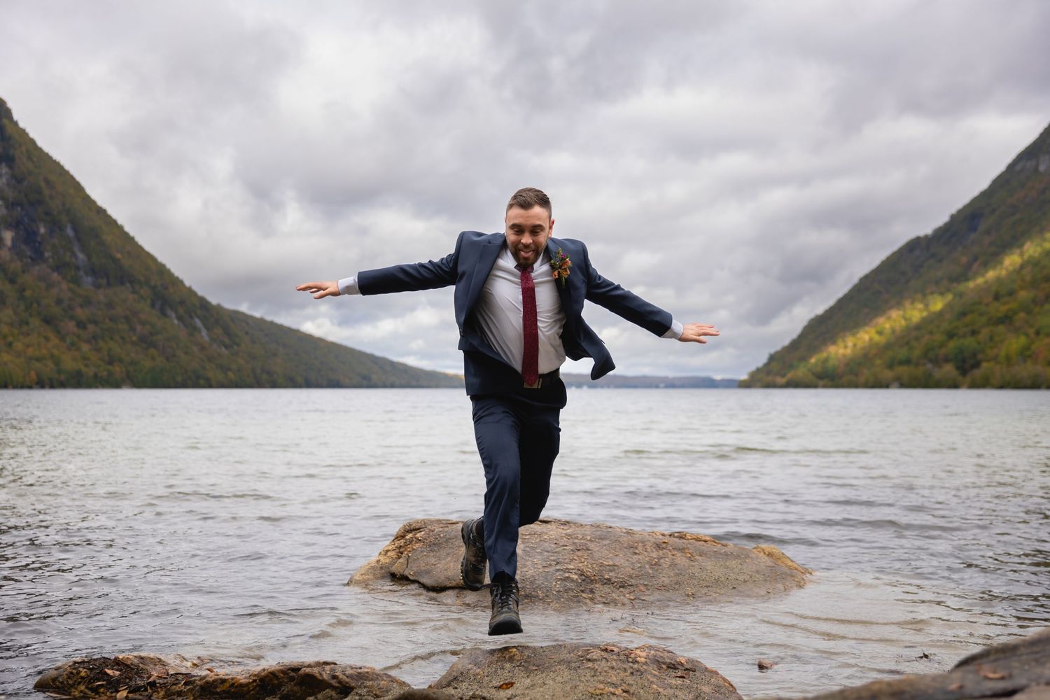 Groom balancing on rock at mountain lake with arms outstretched.