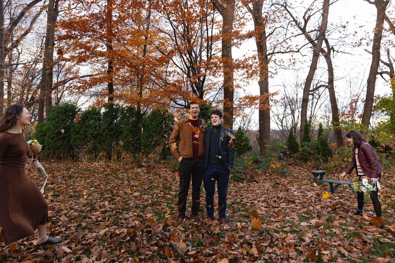 People enjoying an autumn day in the woods with fallen leaves covering the ground and orange foliage on trees.