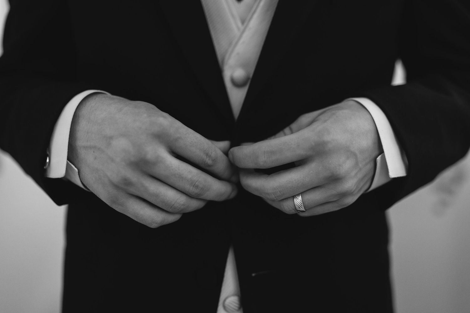 Close-up black and white photo of hands in formal suit adjusting buttons, with a wedding ring visible.