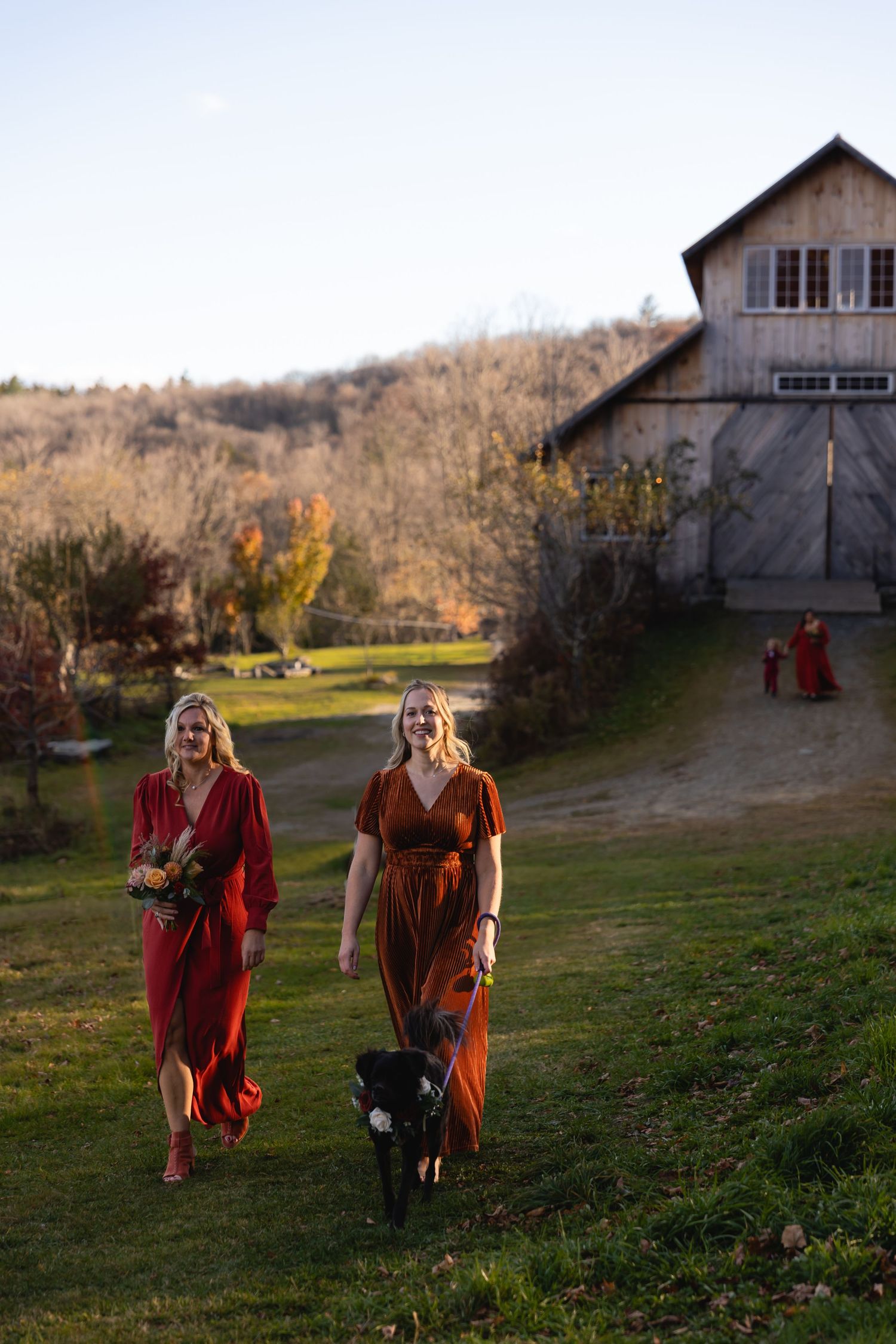 Two people in flowing red and orange dresses walk on a grassy path near a rustic barn at sunset.