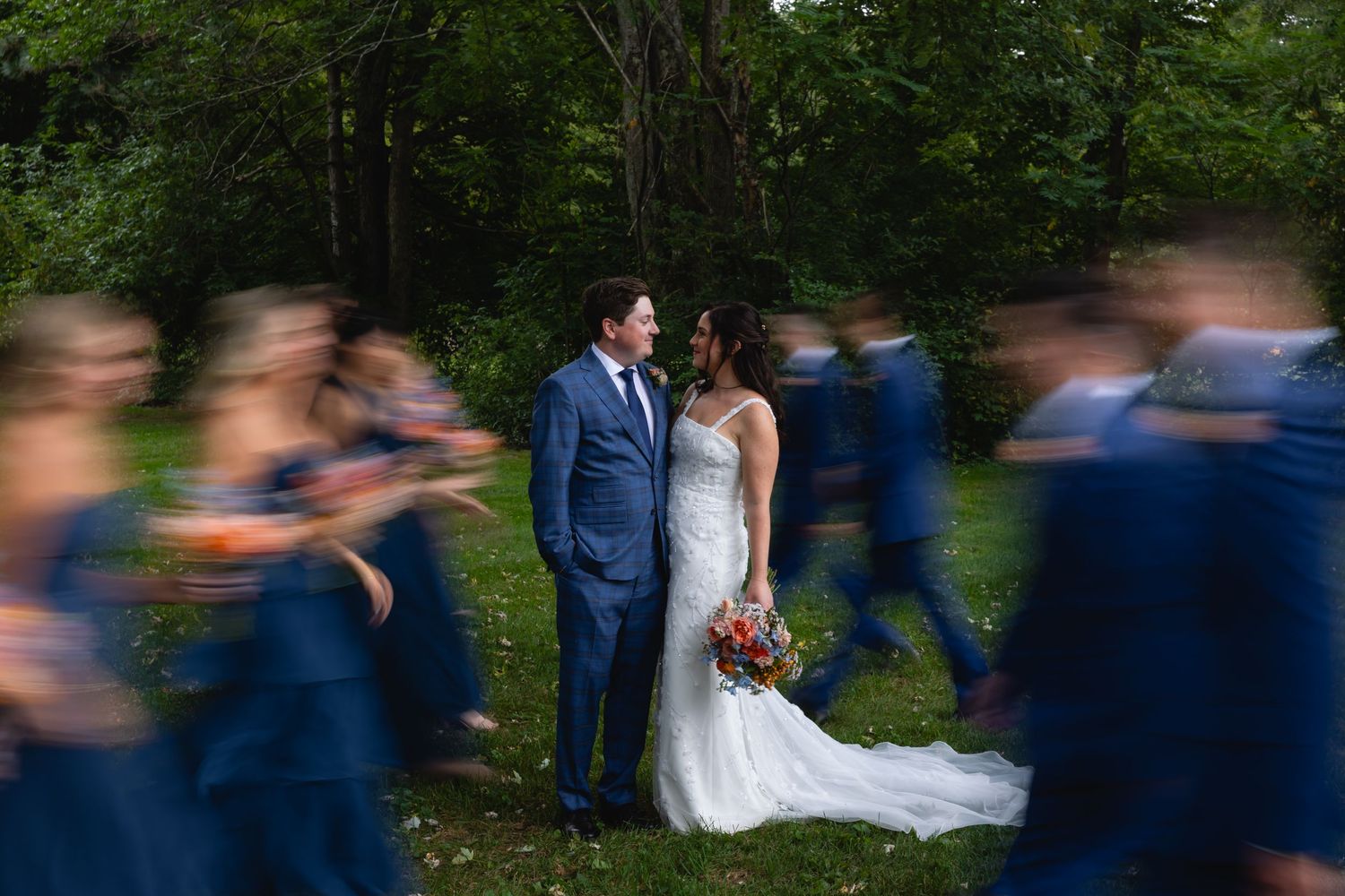 Motion blur captures wedding party movement while couple in white dress and navy suit stay in sharp focus outdoors at forest.