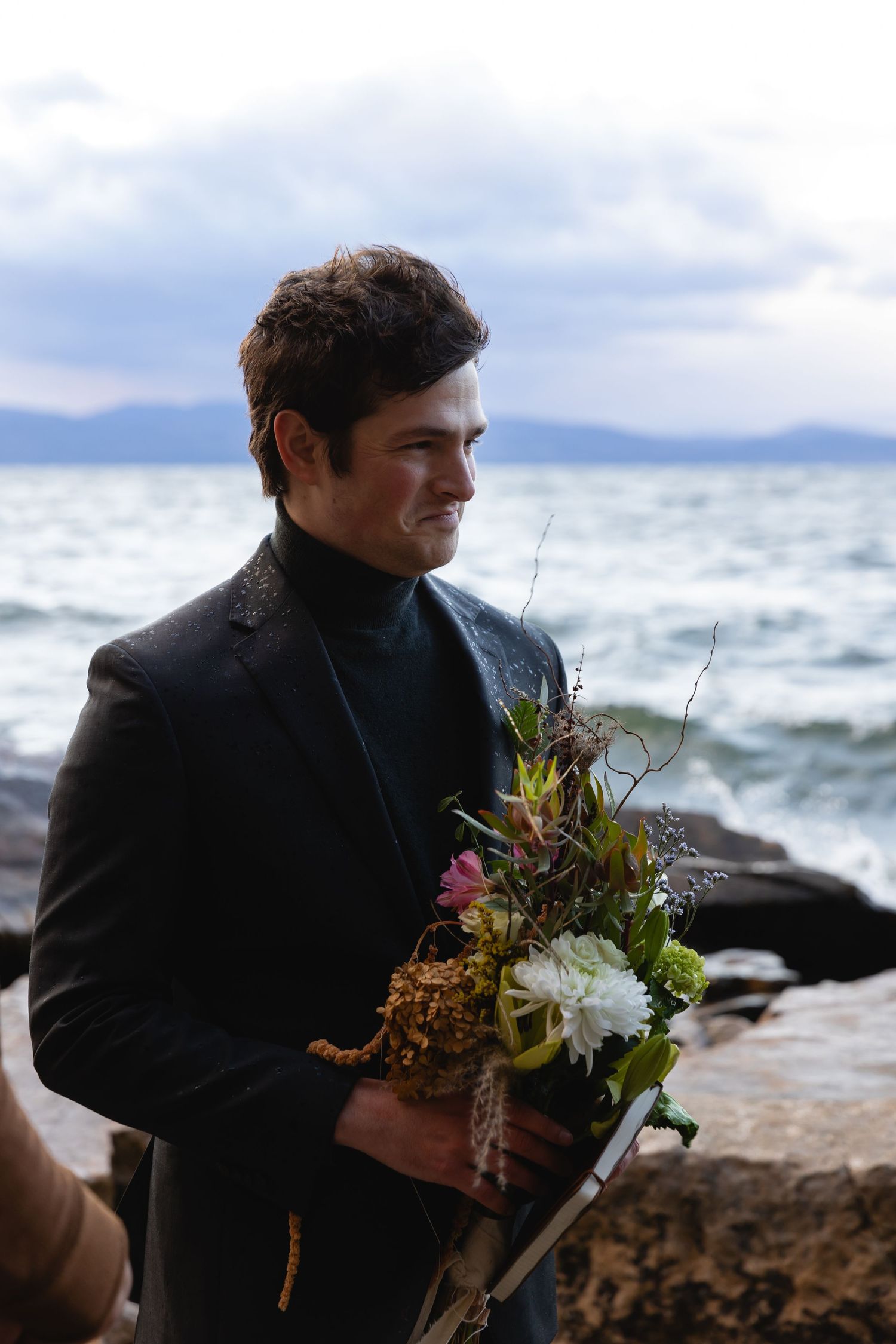 Person in a black turtleneck holds a beautiful wildflower bouquet against a dramatic ocean and mountain backdrop.