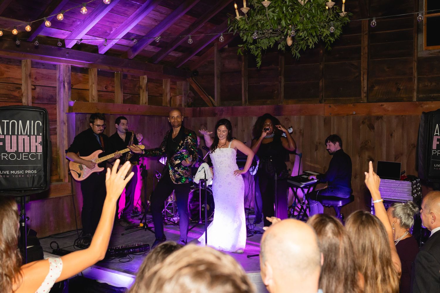 Live band performs on stage at a rustic wedding reception with guests raising their hands in celebration.