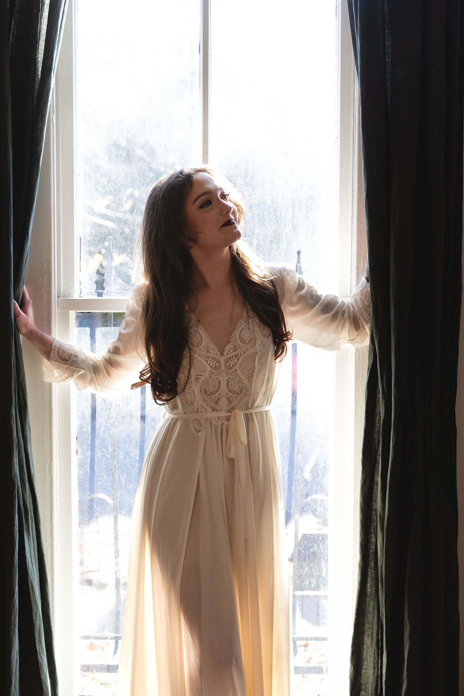 A woman in a flowing white dress stands dramatically between dark curtains at a bright window.