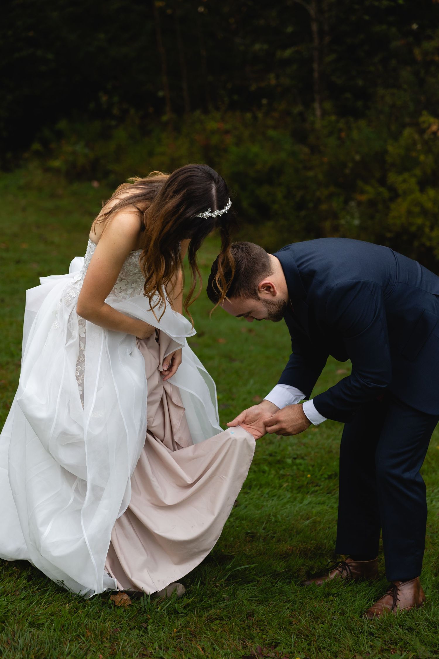 A couple helps adjust wedding attire on a grassy lawn.