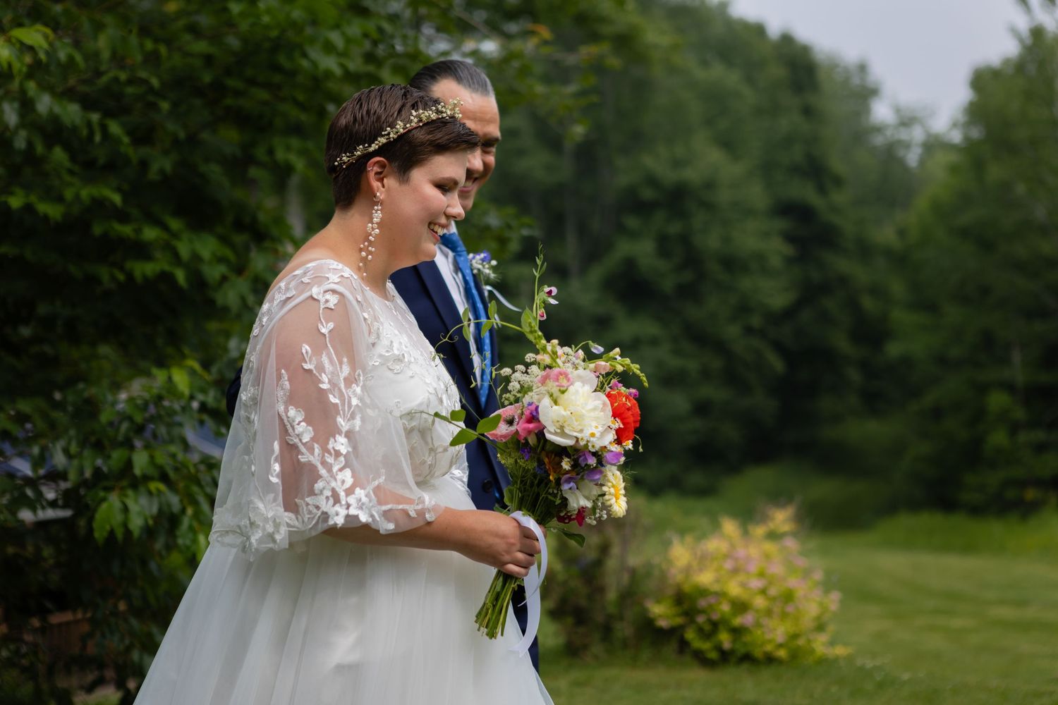 A bride in a lace wedding dress holds a vibrant bouquet in an outdoor garden setting.