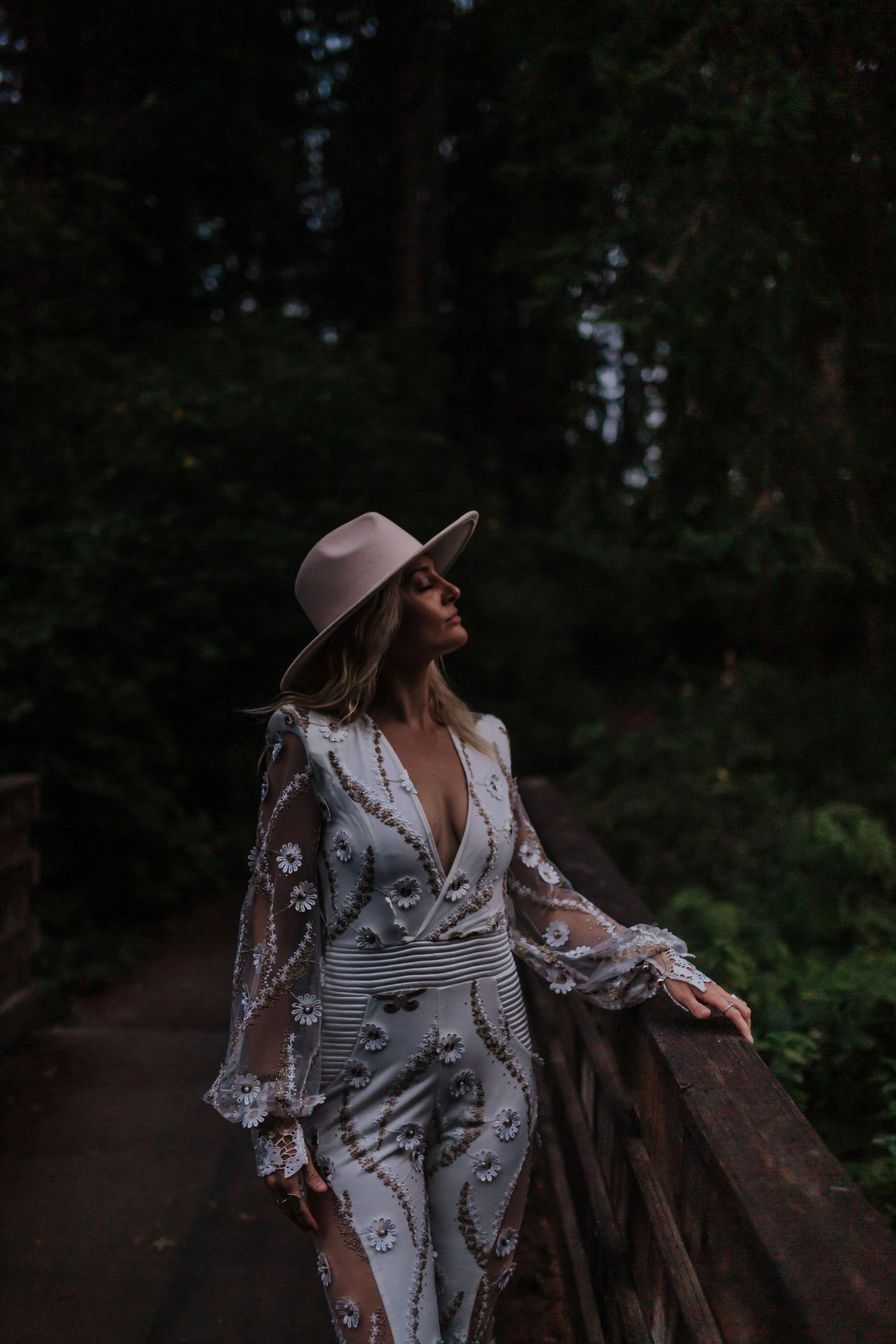 A woman in a white lace dress and hat poses elegantly on a forest path at dusk.