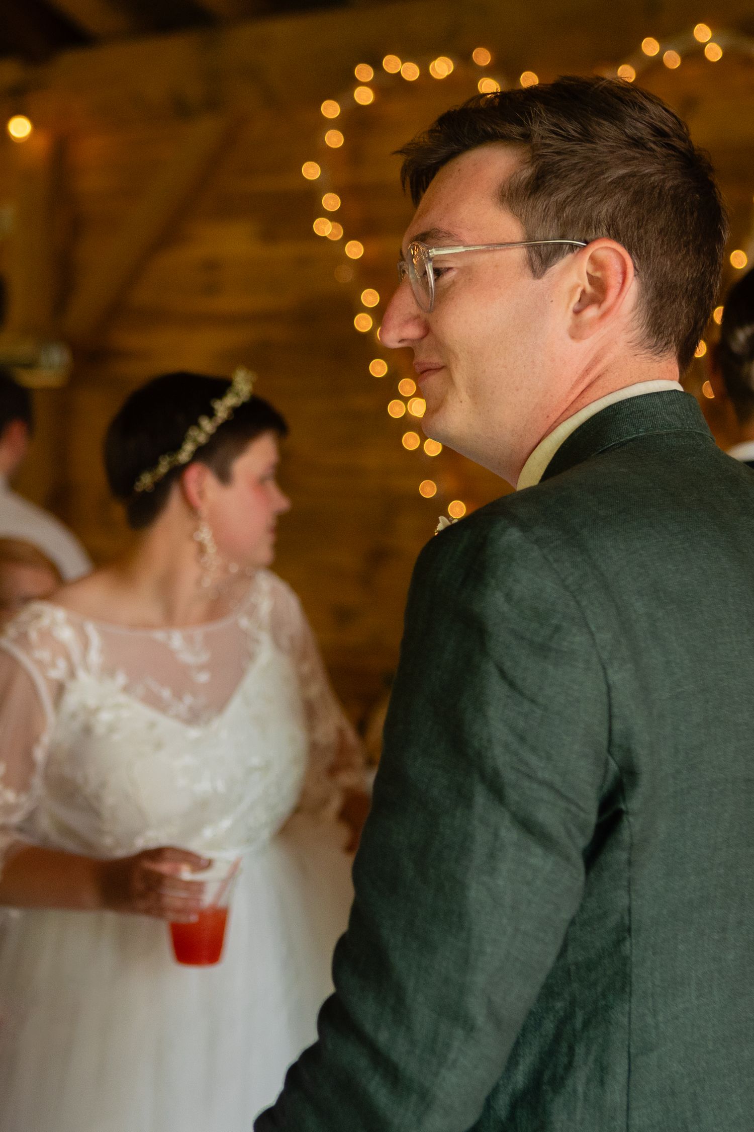 Couple at wedding reception with glowing string lights in background, groom wearing dark suit and glasses.