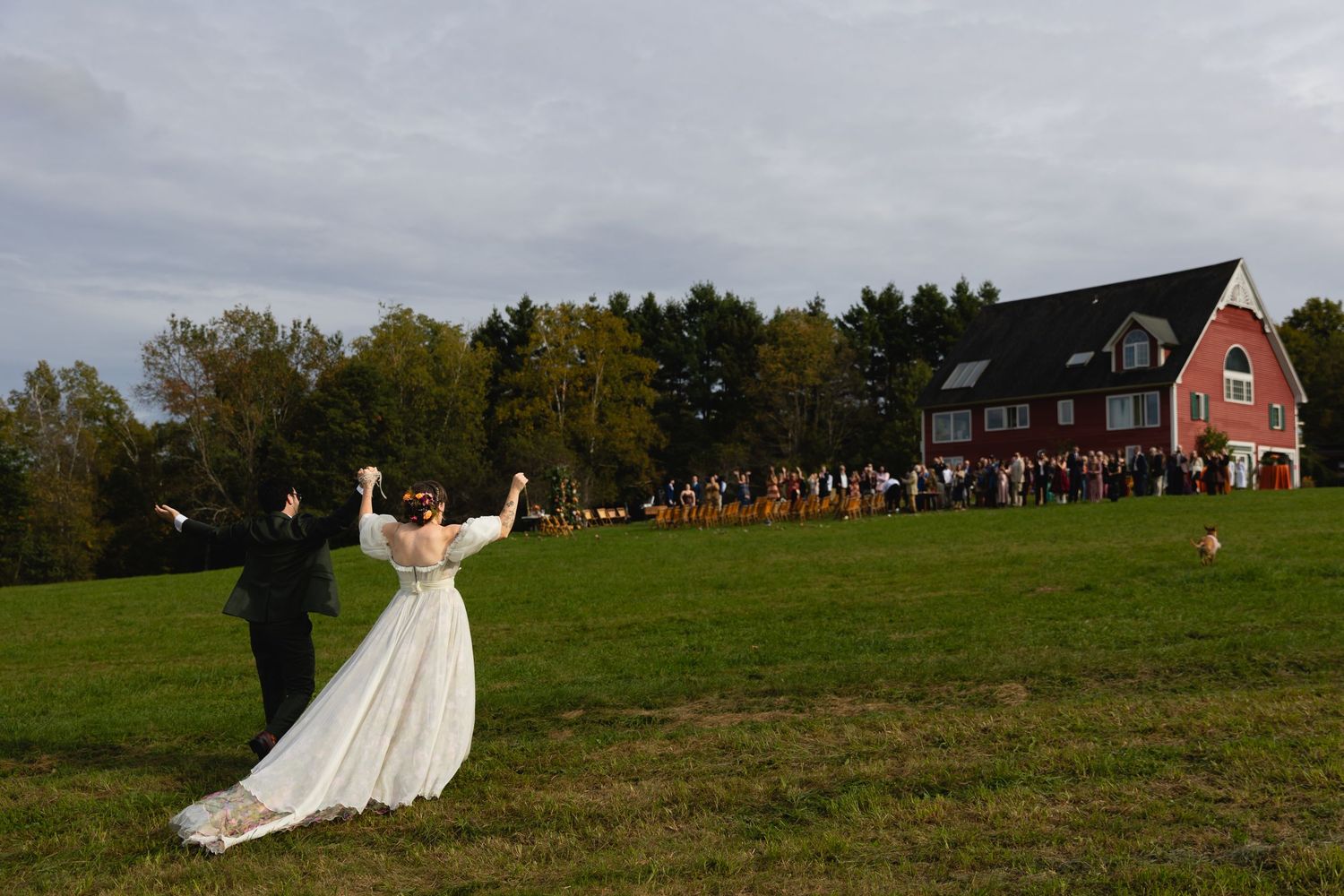 Bride and groom celebrate on green lawn with wedding guests and red barn in background on autumn day.