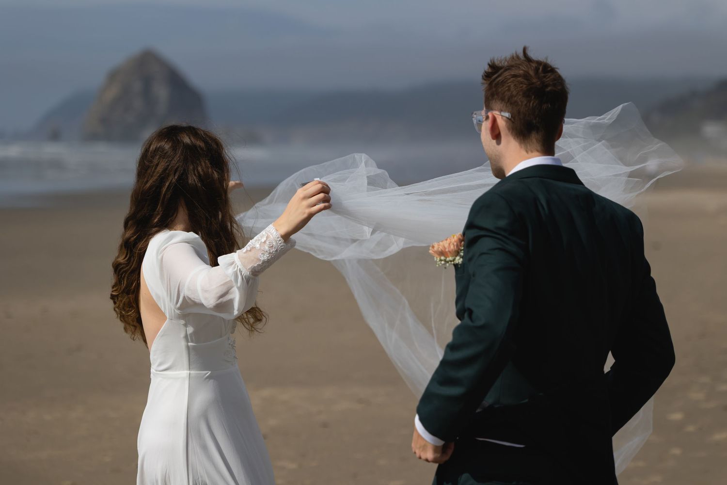 A couple in wedding attire walks along a misty beach while the bride's veil flows dramatically in the wind.