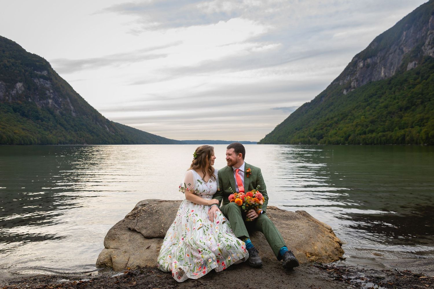 A couple sits on a rock by a serene mountain lake surrounded by towering peaks at sunset.