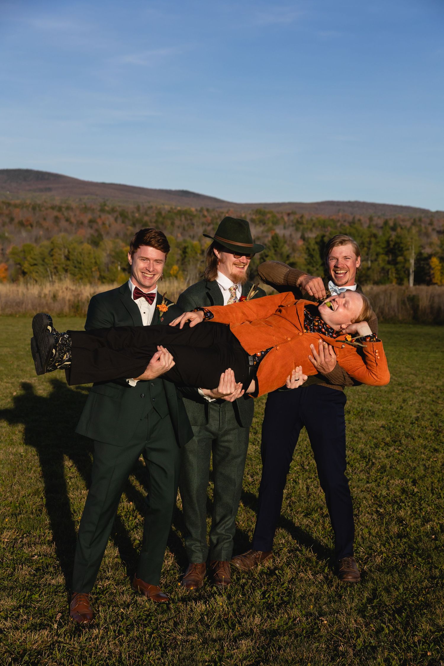 Three people in formal attire playfully pose outdoors against an autumn mountain landscape on a sunny day.