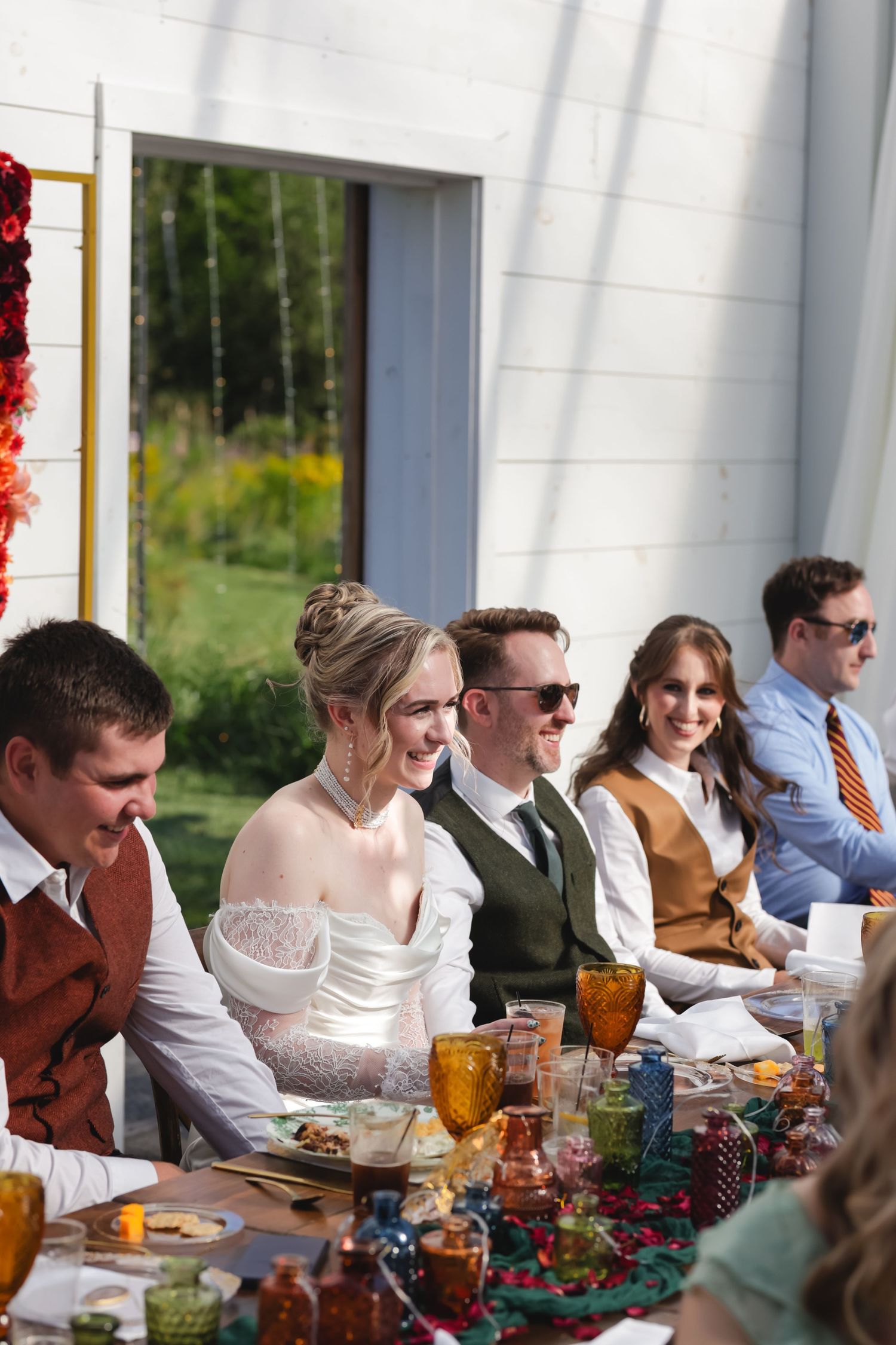 Wedding guests enjoy a meal at a long table during an outdoor reception with white columns and greenery in view.