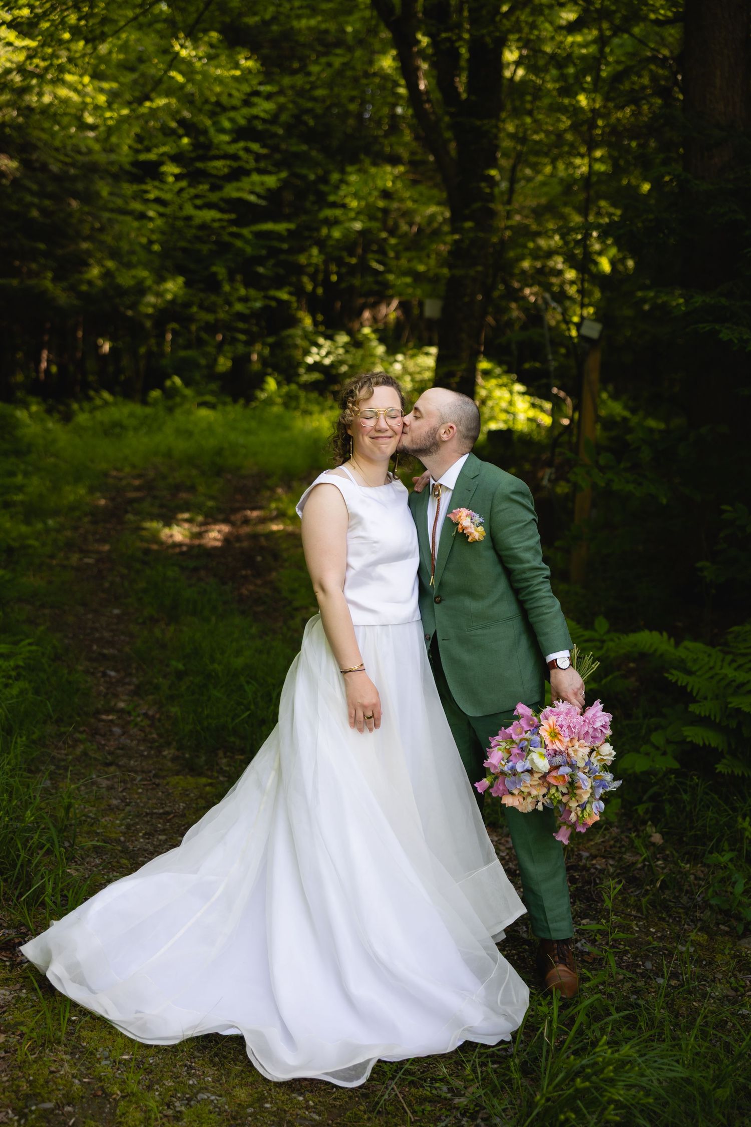 A couple in wedding attire embraces in a lush green forest setting with a colorful bouquet of flowers.