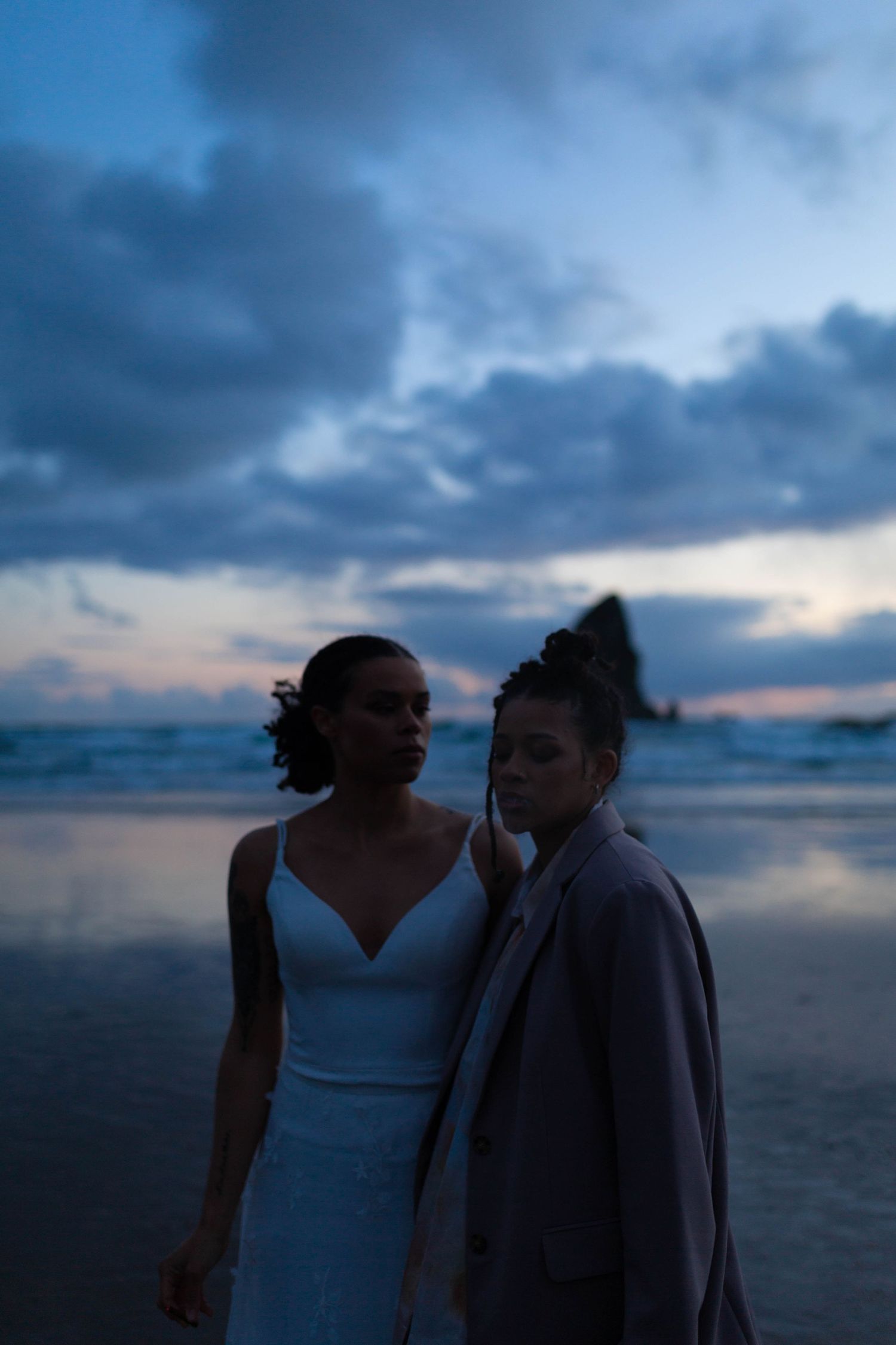 Two people share a tender moment on a beach against a dramatic twilight sky.