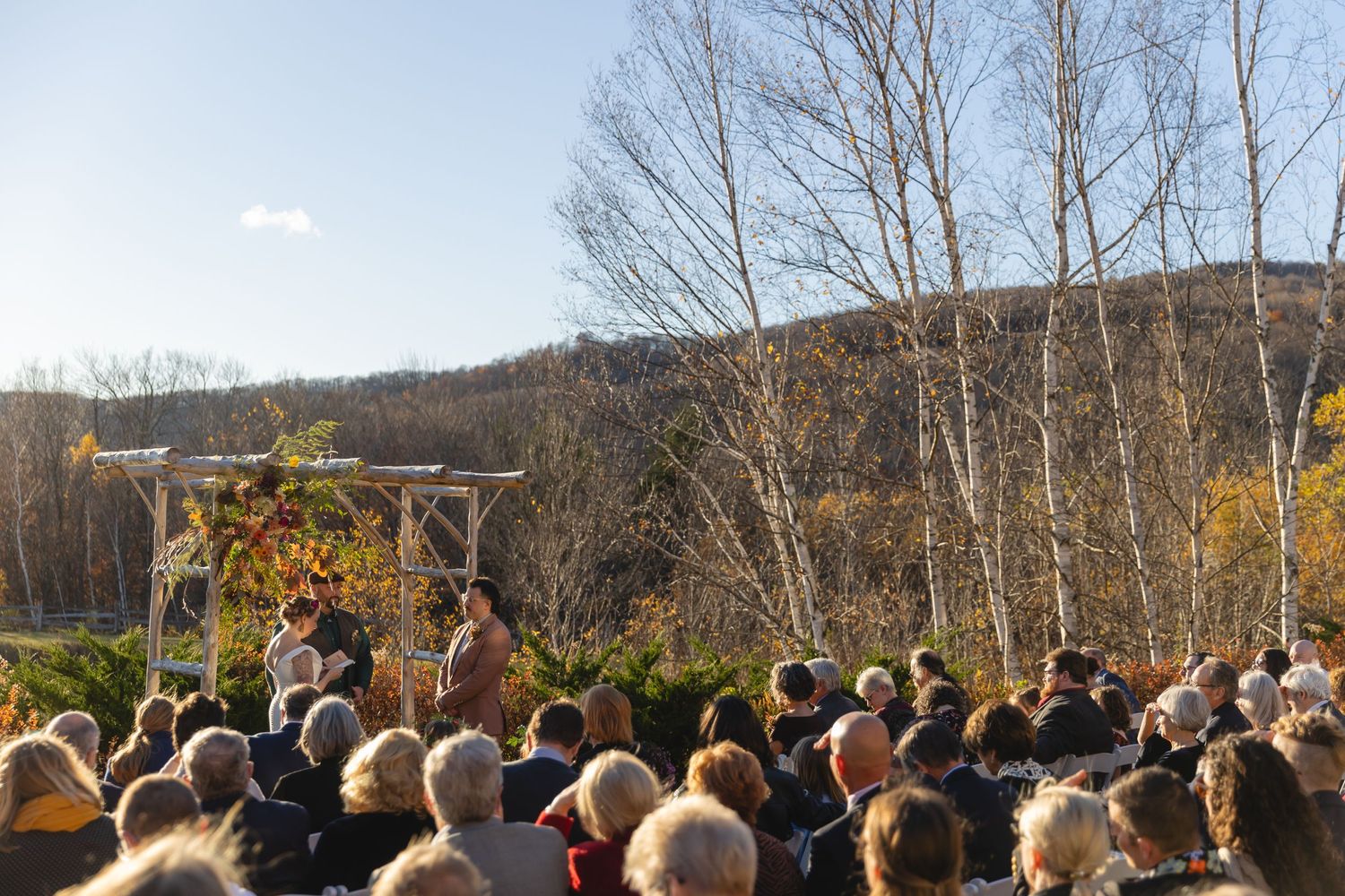 A large crowd watches an outdoor autumn wedding ceremony beneath a decorated wooden arch.
