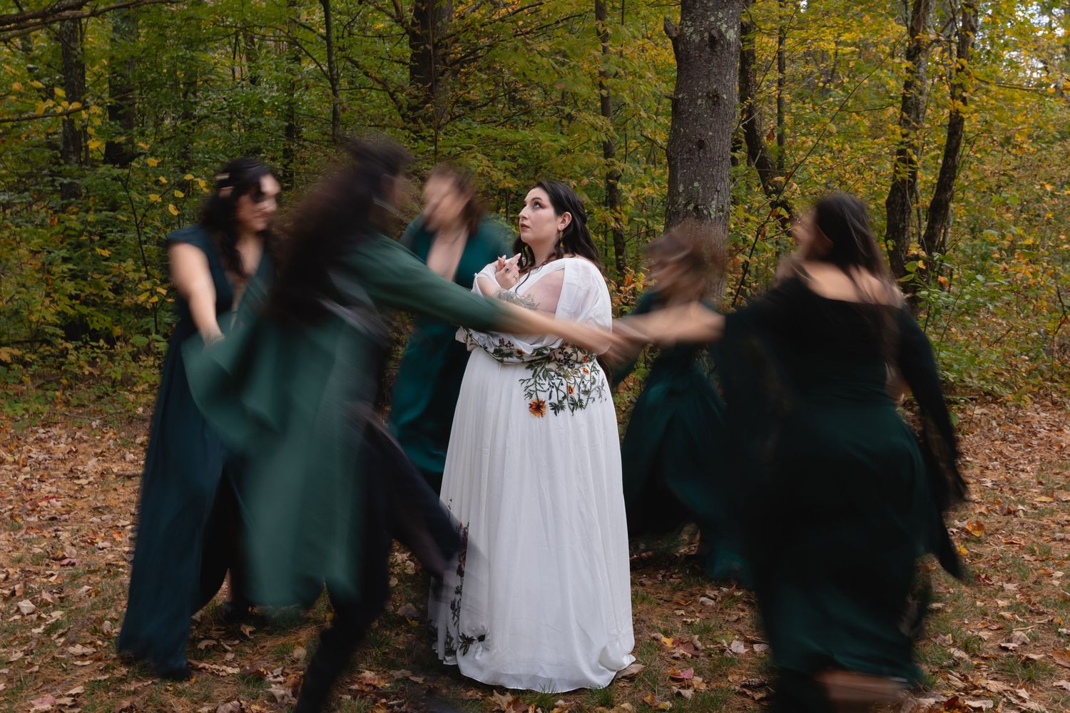 Bride in white dress surrounded by bridesmaids in green dresses dancing in forest.