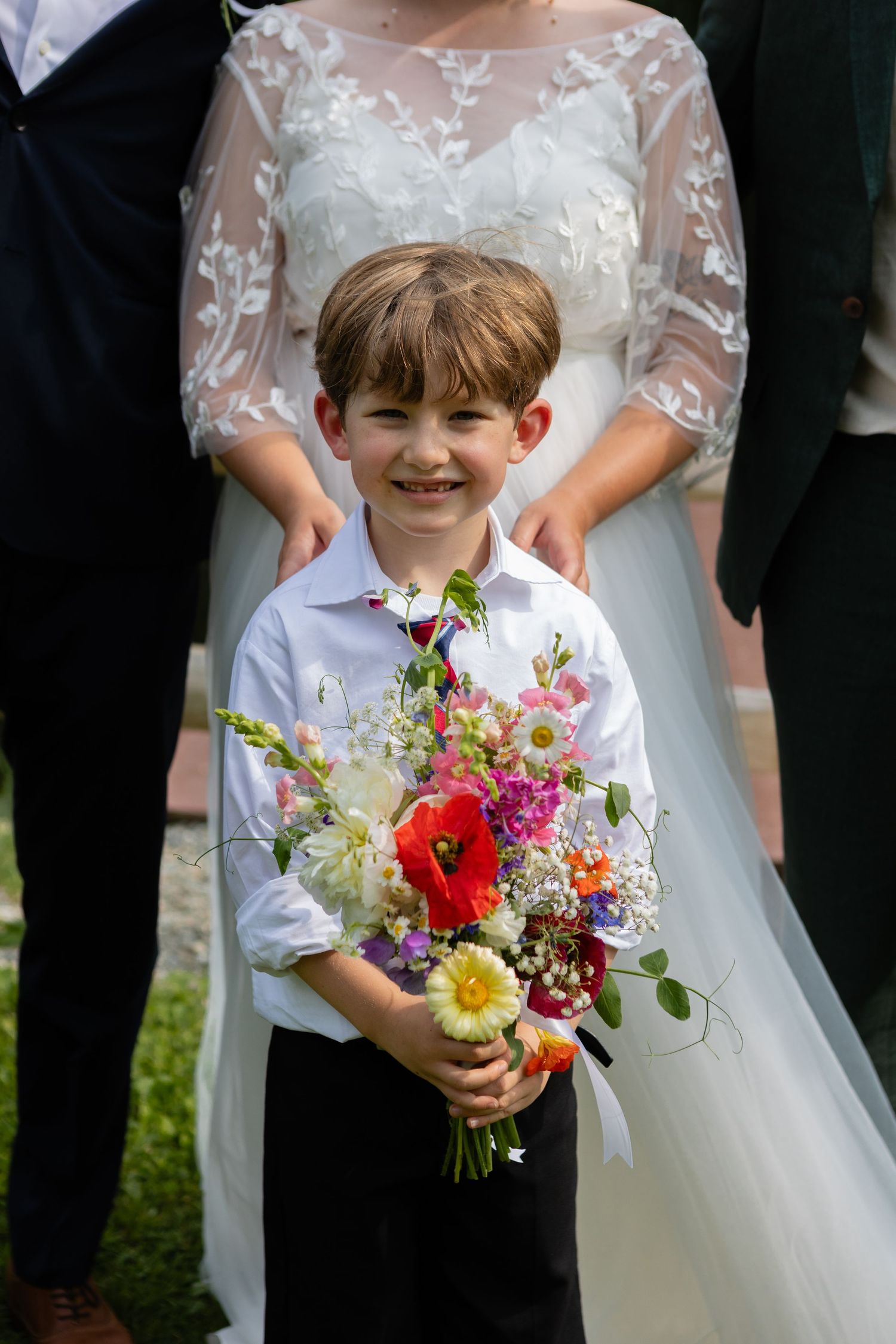 A colorfully dressed child holds a vibrant wildflower bouquet in front of a wedding dress at an outdoor ceremony.