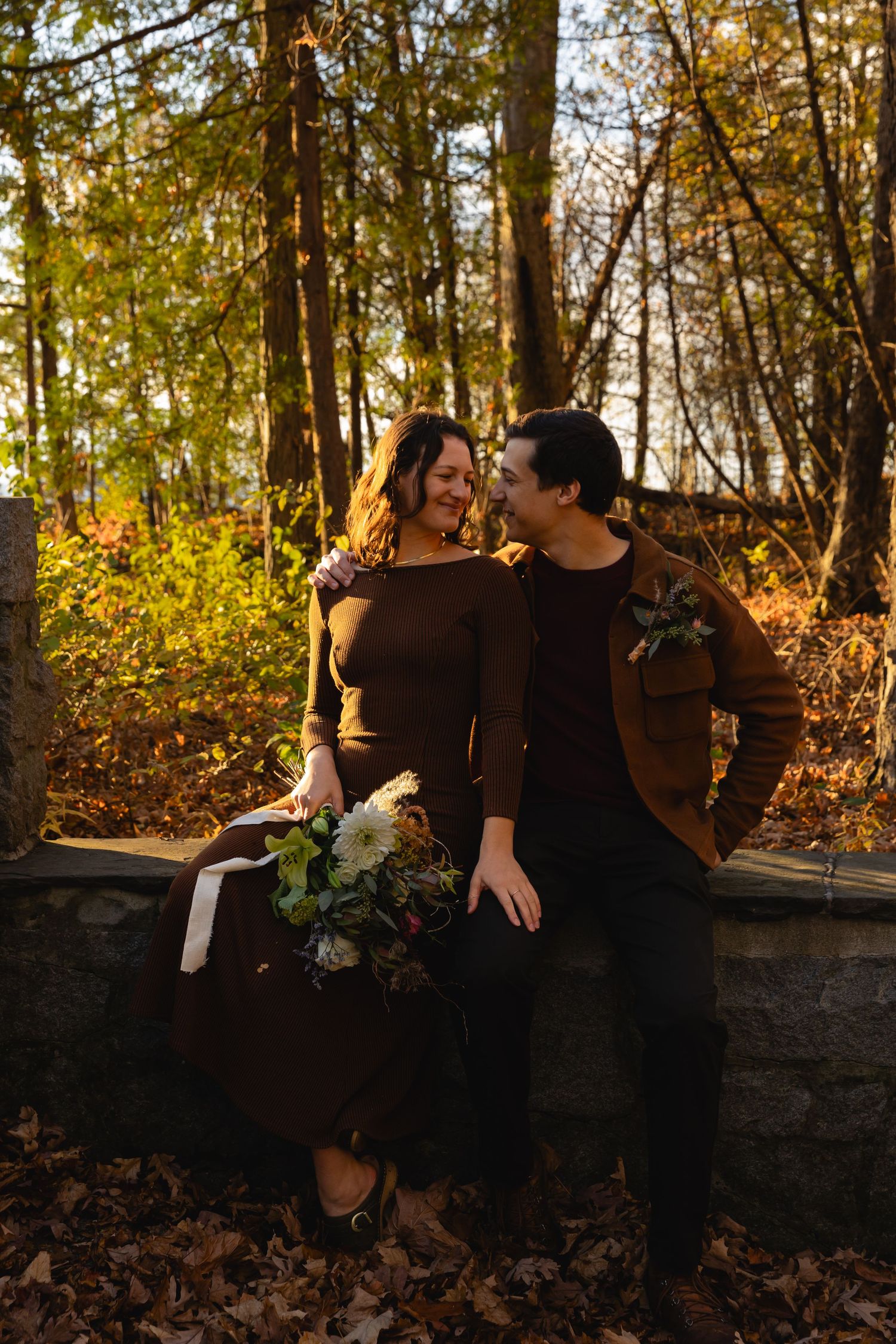A couple shares a romantic moment on a stone wall surrounded by autumn trees in warm golden light.