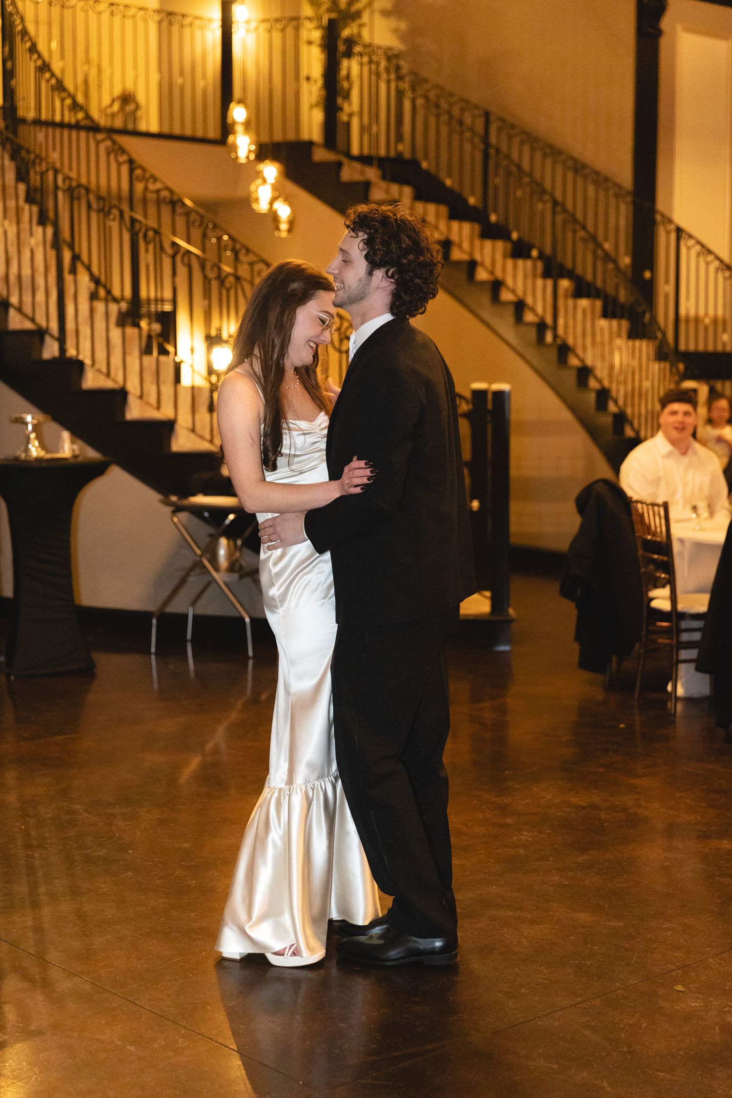 A couple shares an intimate dance moment in a grand ballroom with elegant staircase and warm lighting in the background.