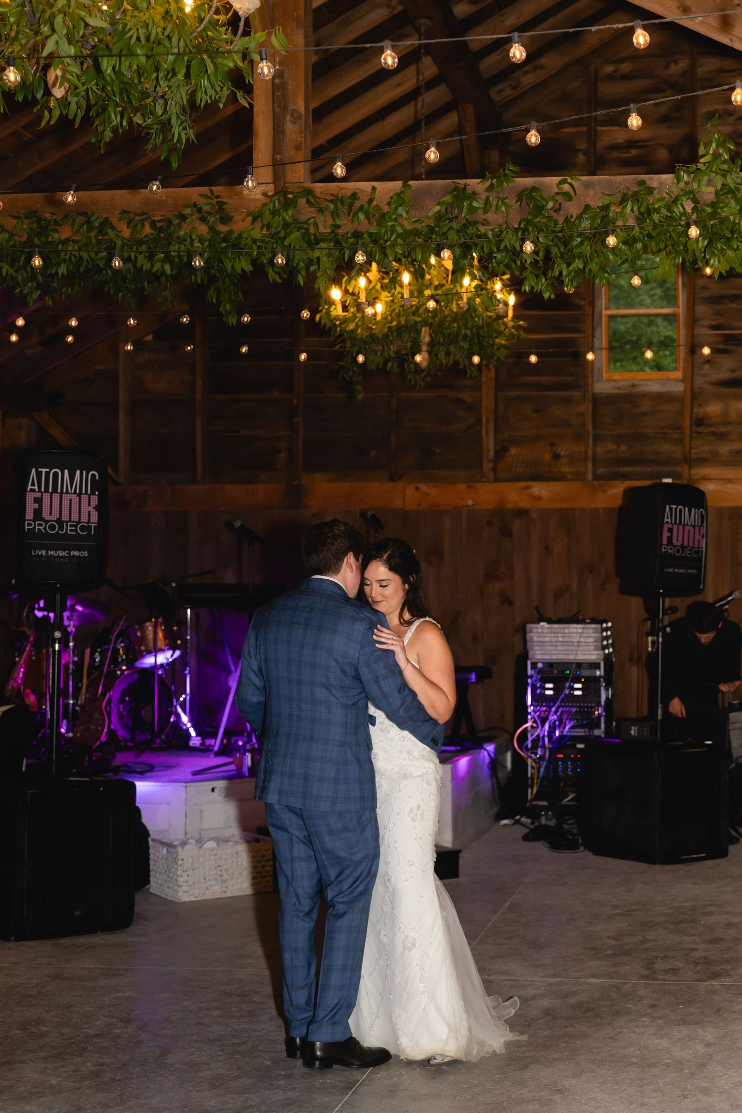 A couple shares their first dance in a rustic barn venue decorated with string lights and greenery.