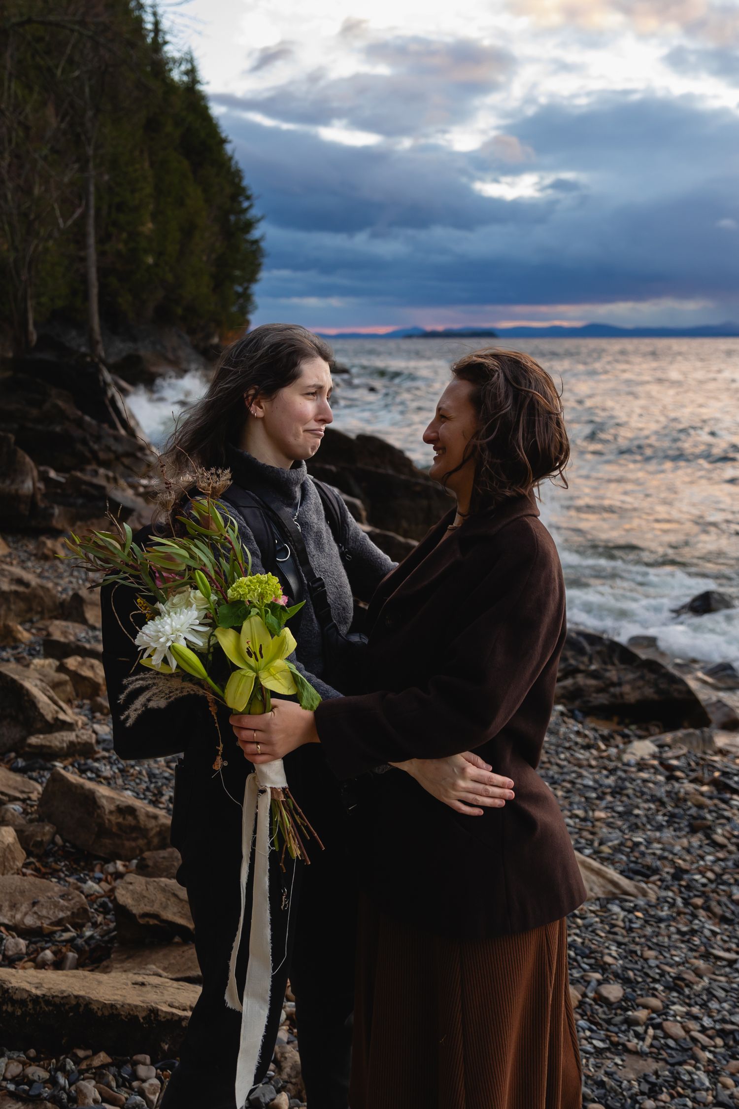 Two people stand on a rocky coastline at sunset holding yellow flowers with dramatic stormy clouds overhead.
