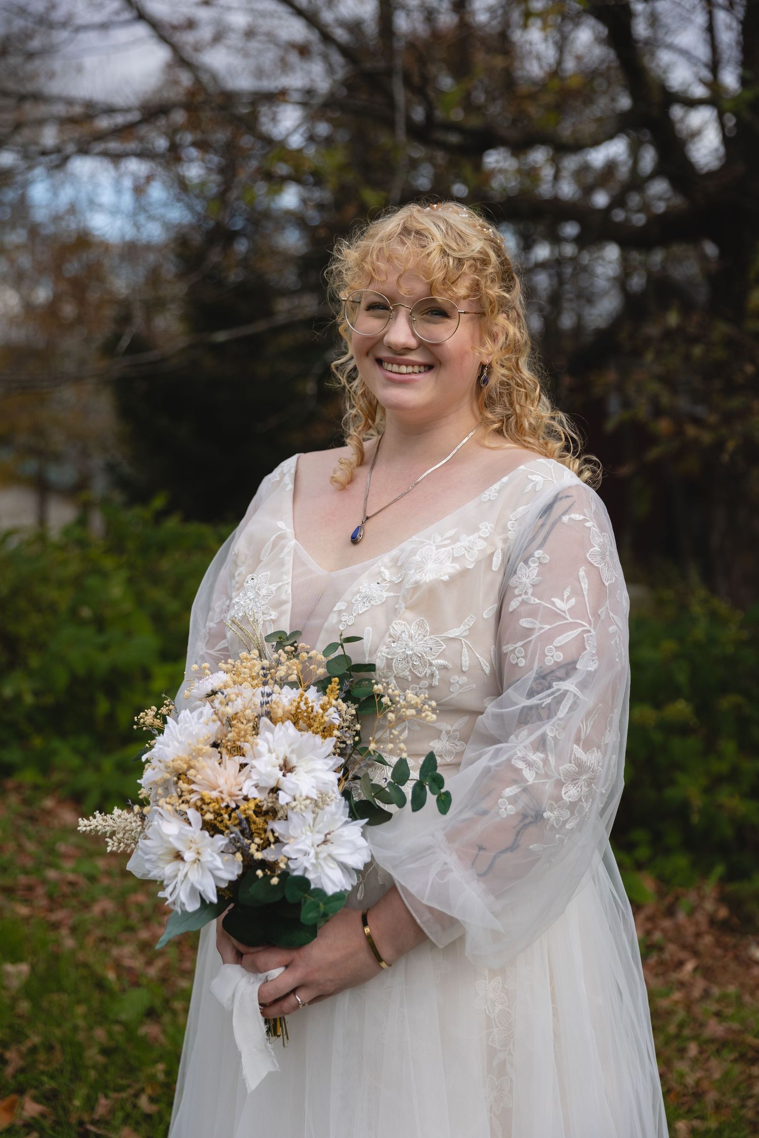 A bride smiling while holding a bouquet of white and yellow flowers in an autumn garden.