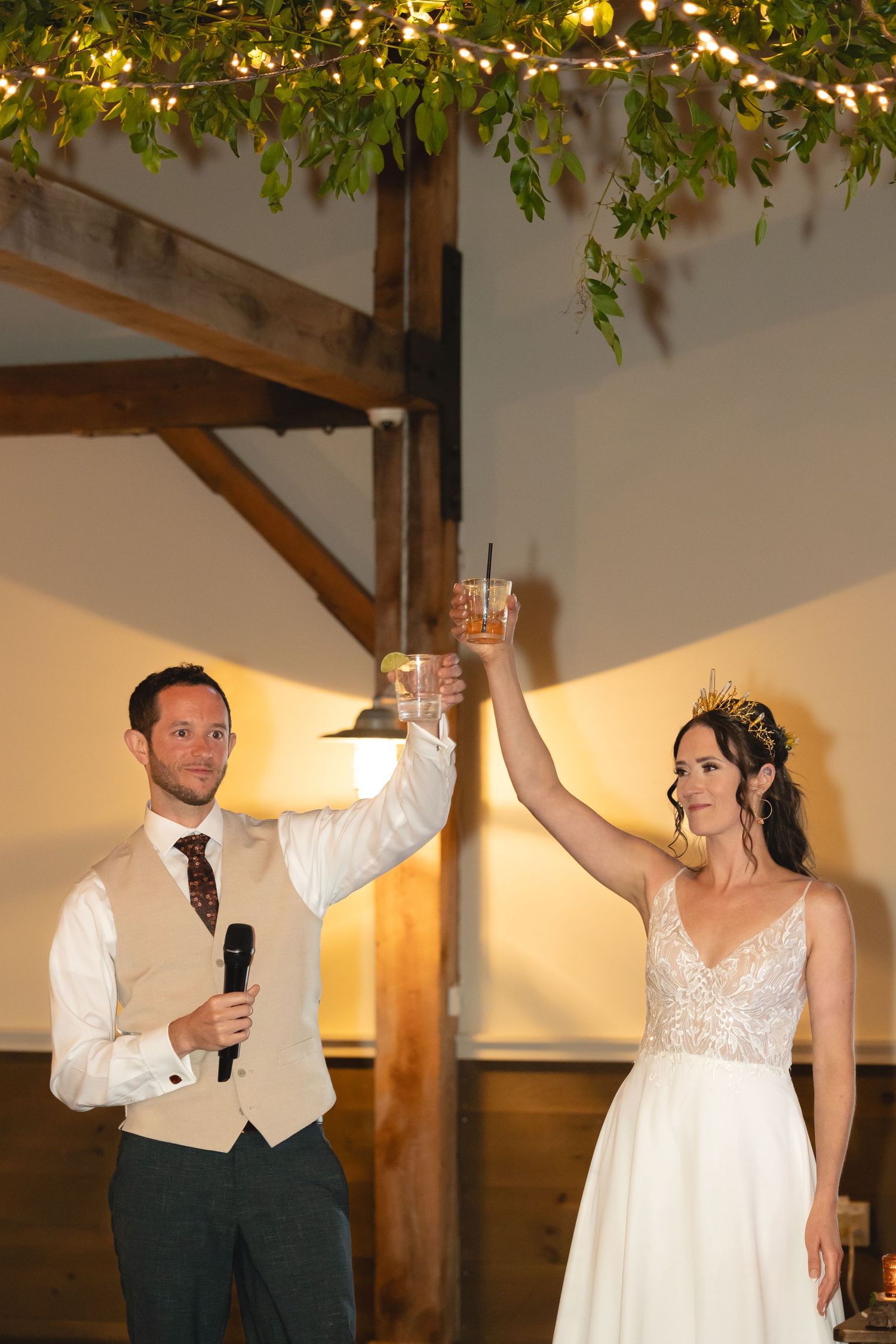 Wedding couple raising glasses in celebratory toast under greenery and lights.
