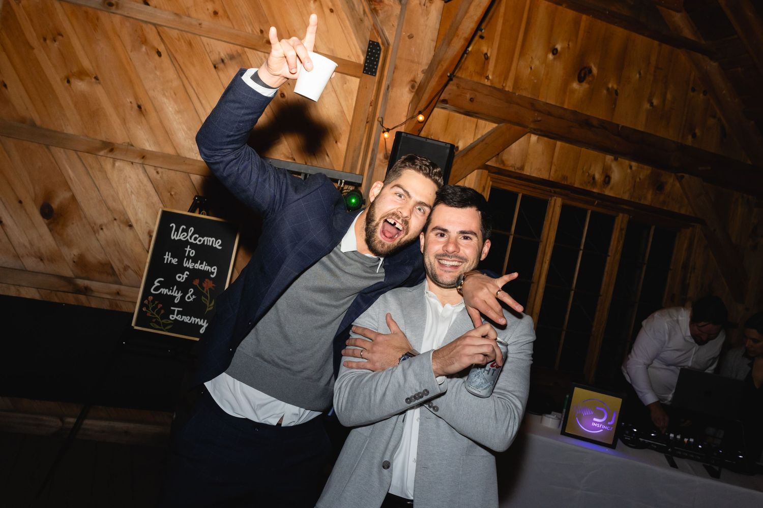 Two energetic party guests strike playful poses on the dance floor at a rustic wooden venue during a celebration.