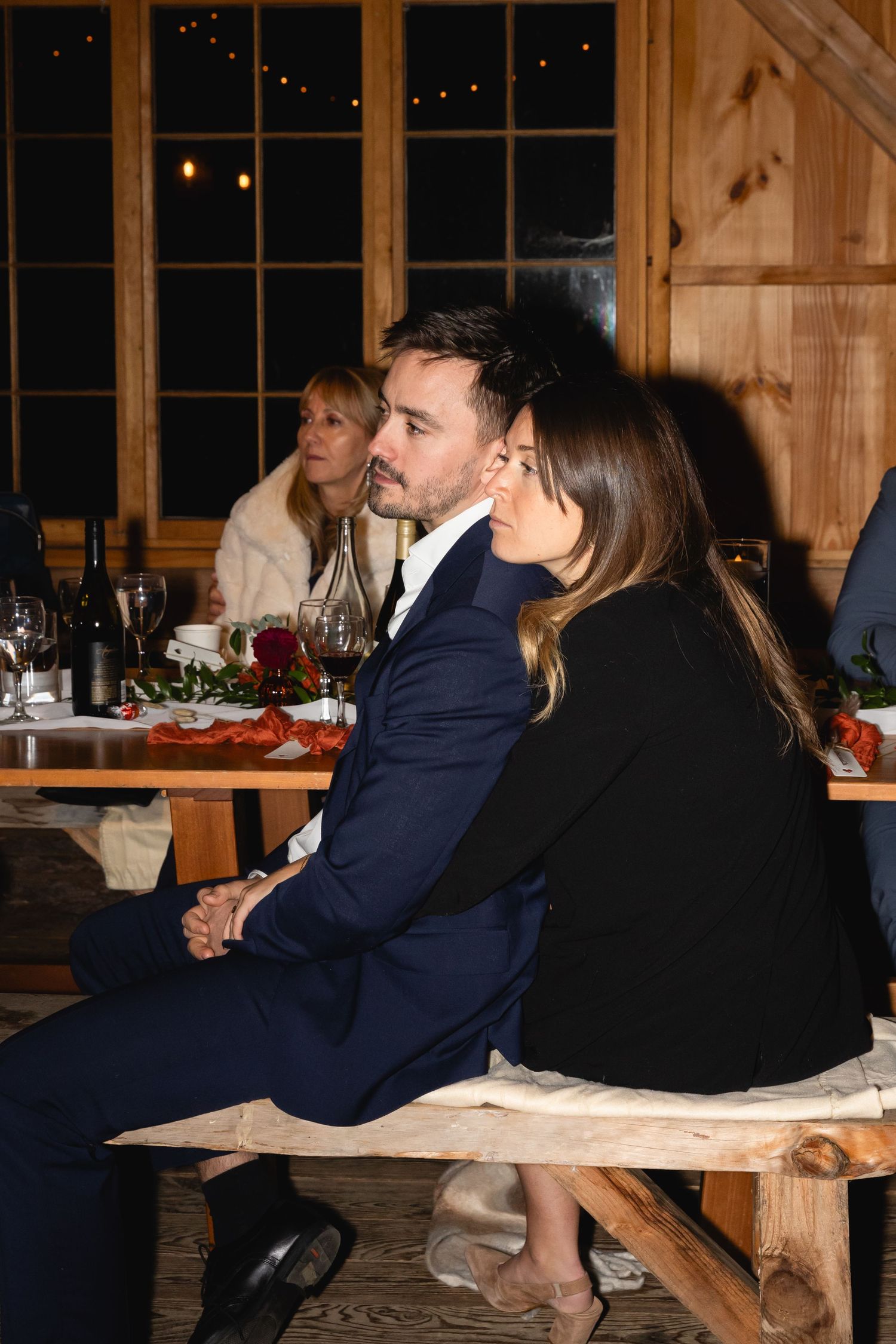 Couple sitting together at rustic wooden table during evening dinner gathering with wine bottles and plates.