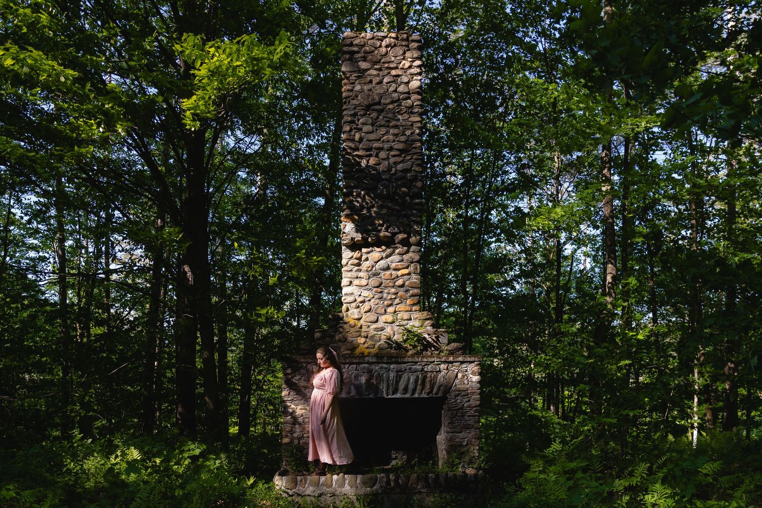 Woman in pink dress stands beside stone chimney surrounded by lush green forest trees.