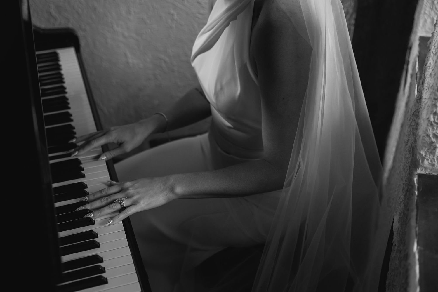 Black and white close-up of a bride in a veil playing piano keys with graceful hands on her wedding day.