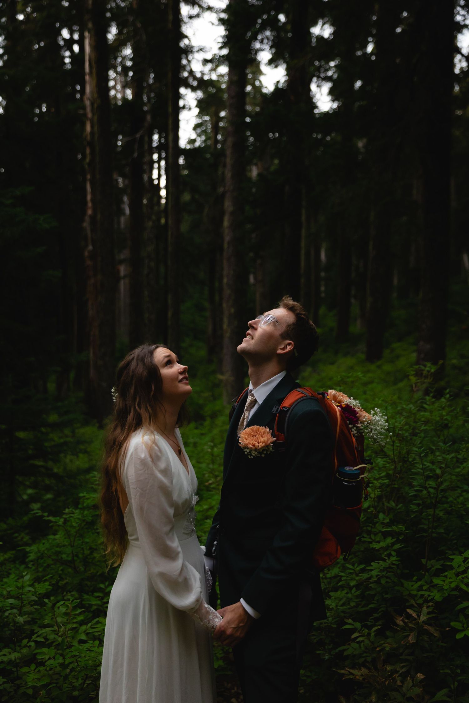 Bride and groom hold hands laughing in a lush dark forest, groom wearing a boutonniere and carrying a backpack.