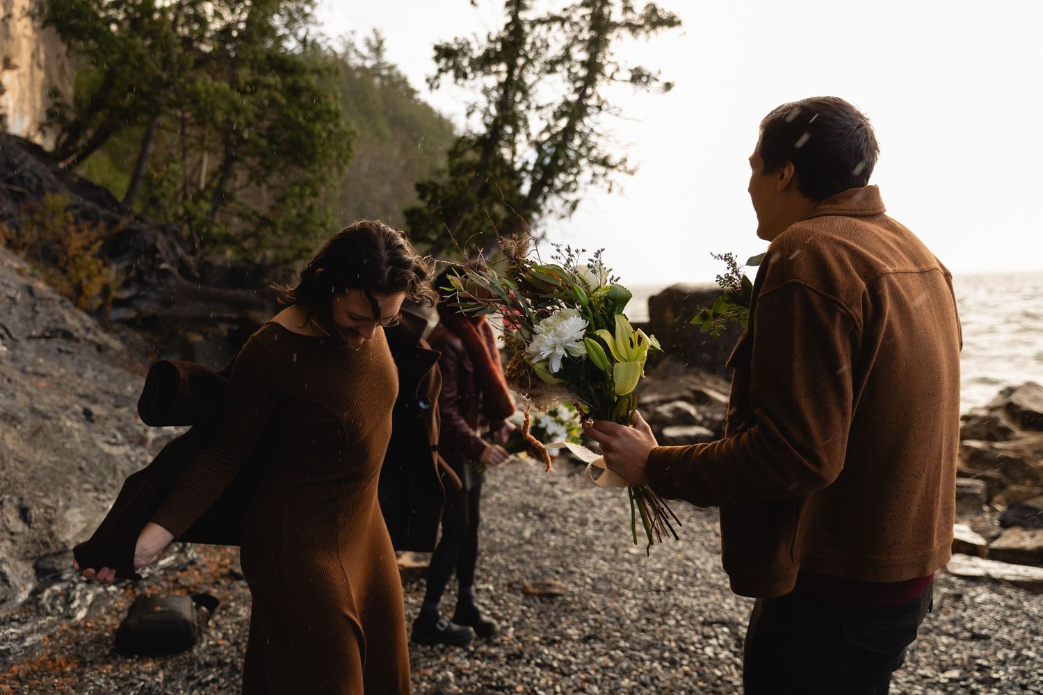 Man presenting a white floral bouquet to a woman on a rocky beach near cliffs at sunset.