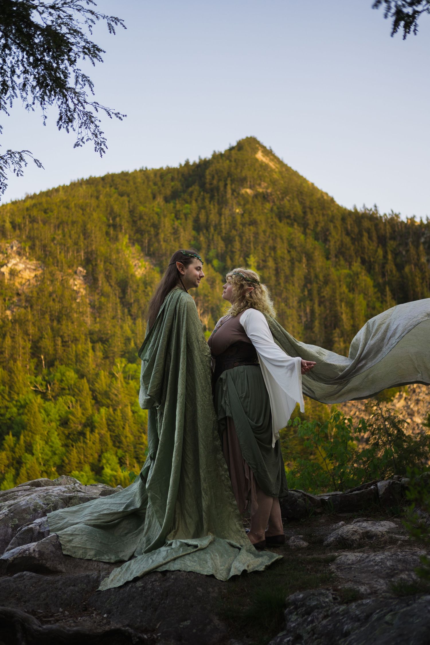 Two women in medieval elven gowns facing each other on rocks with mountain backdrop.