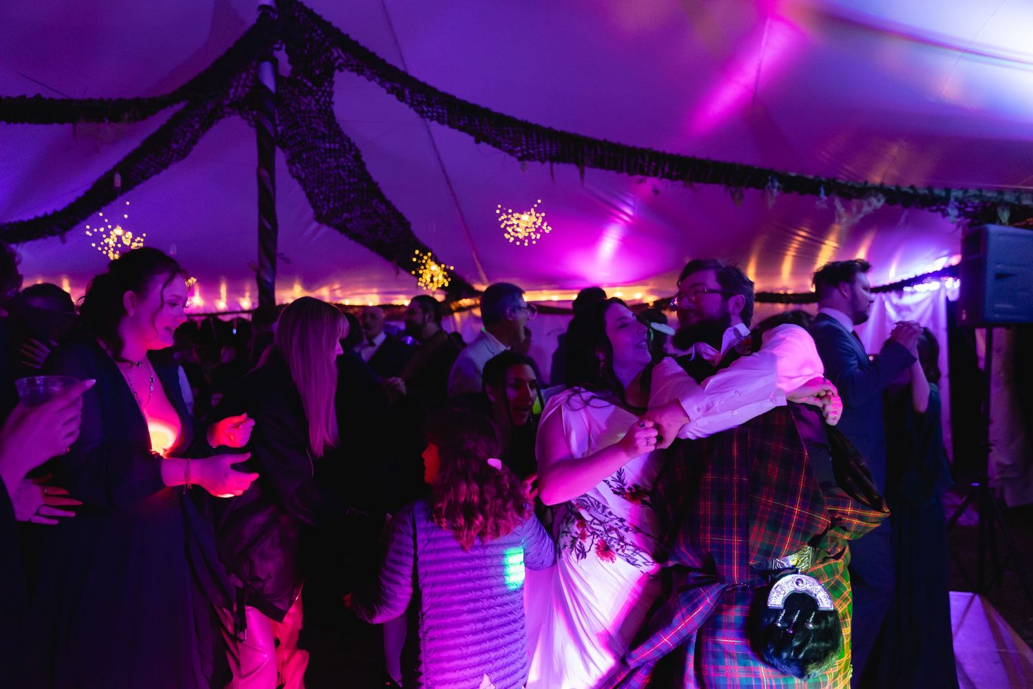 Wedding guests dancing under a decorated tent with purple and pink lighting.