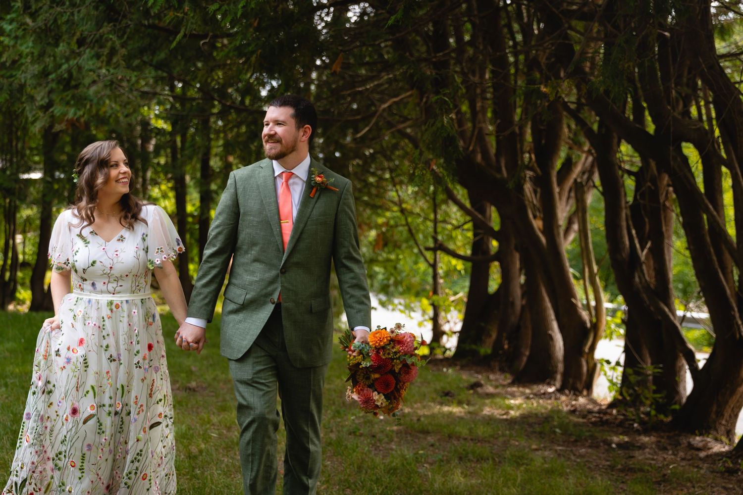 A couple walks together in a wooded outdoor setting wearing wedding attire with sunlight filtering through trees.