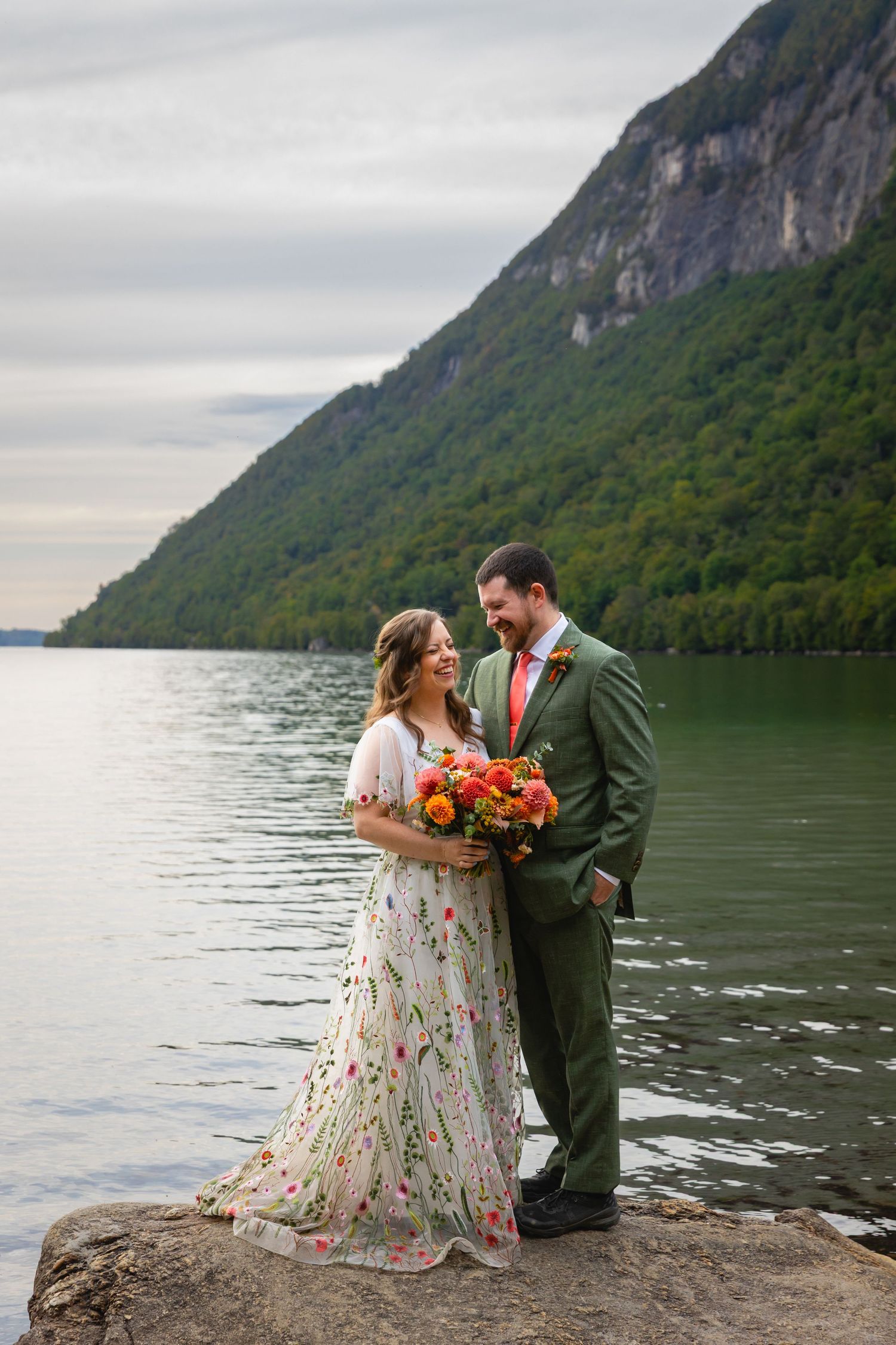 A couple shares a romantic moment on a rocky shore with mountains and a lake creating a stunning backdrop.